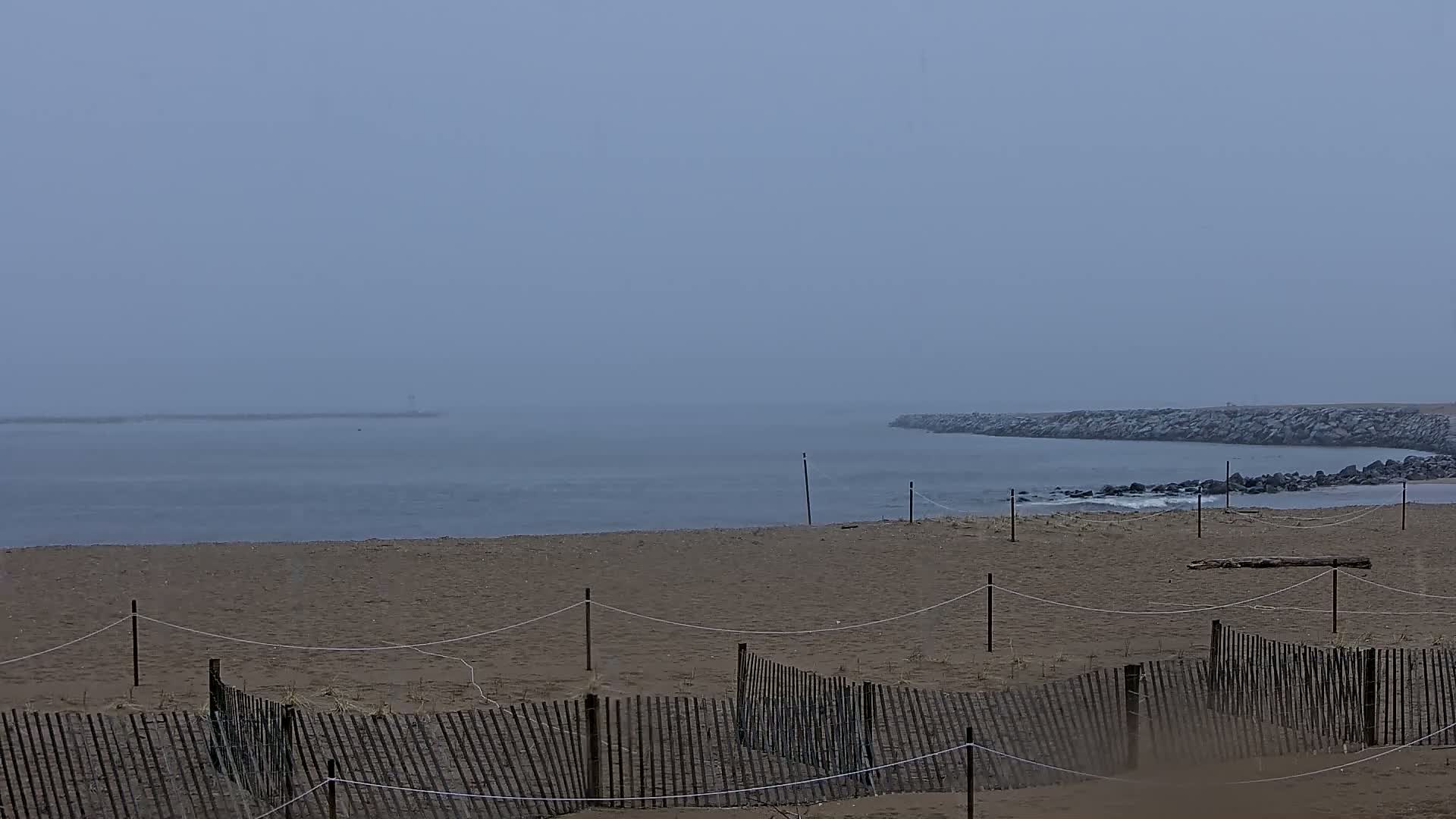 A sandy beach with a wooden fence is visible, under a foggy sky, overlooking a calm body of water with a rock jetty in the distance.