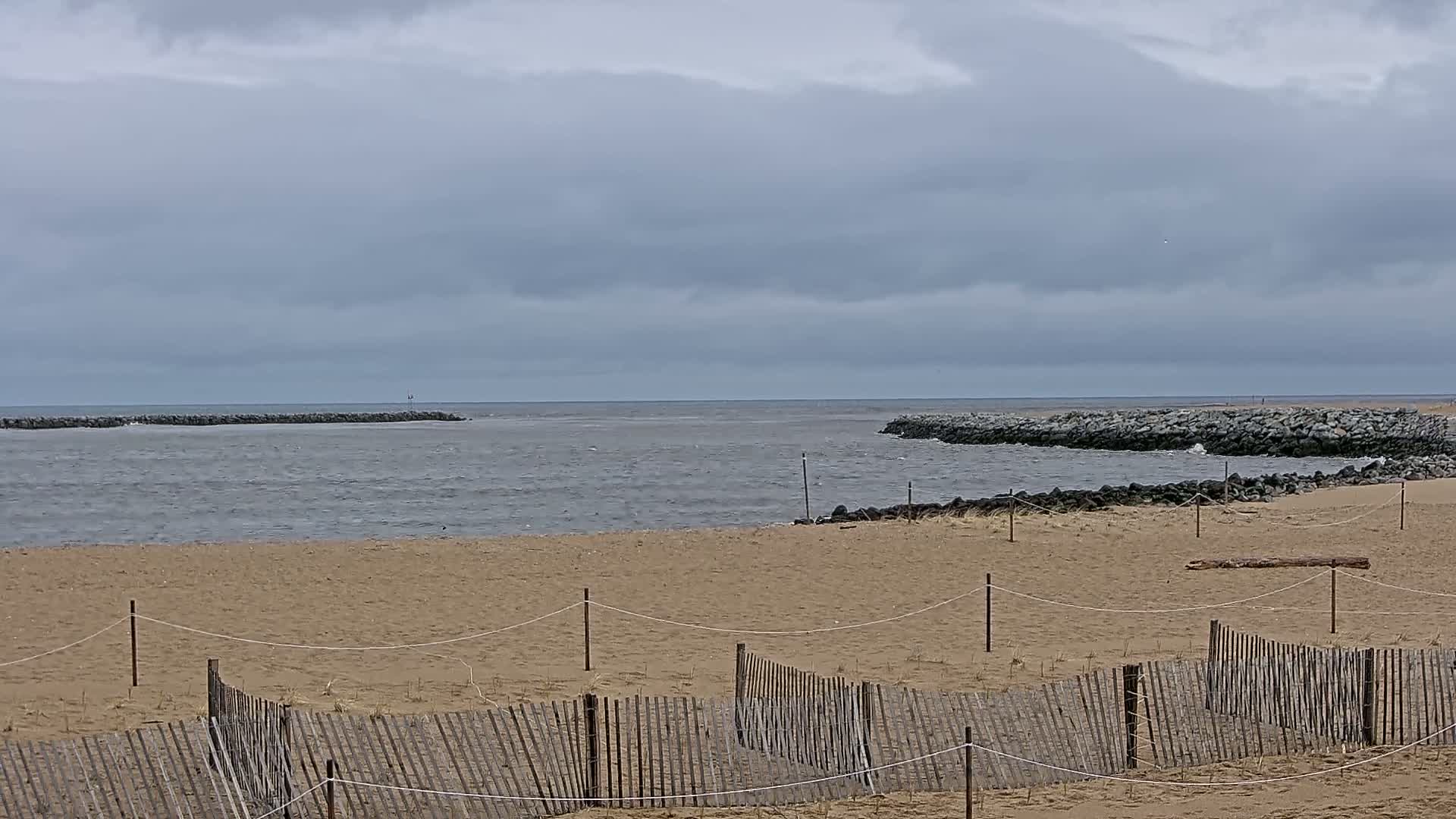A sandy beach with a low wooden fence stretches toward a calm, gray ocean under a cloudy sky, with rock jetties visible in the distance.