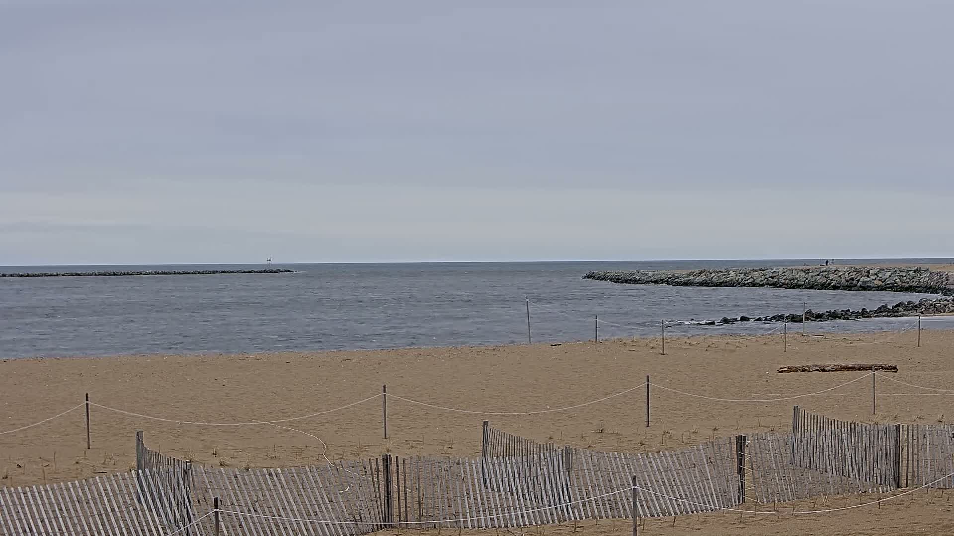 A sandy beach with a wooden fence is seen in front of a calm, gray ocean with rock jetties on either side.