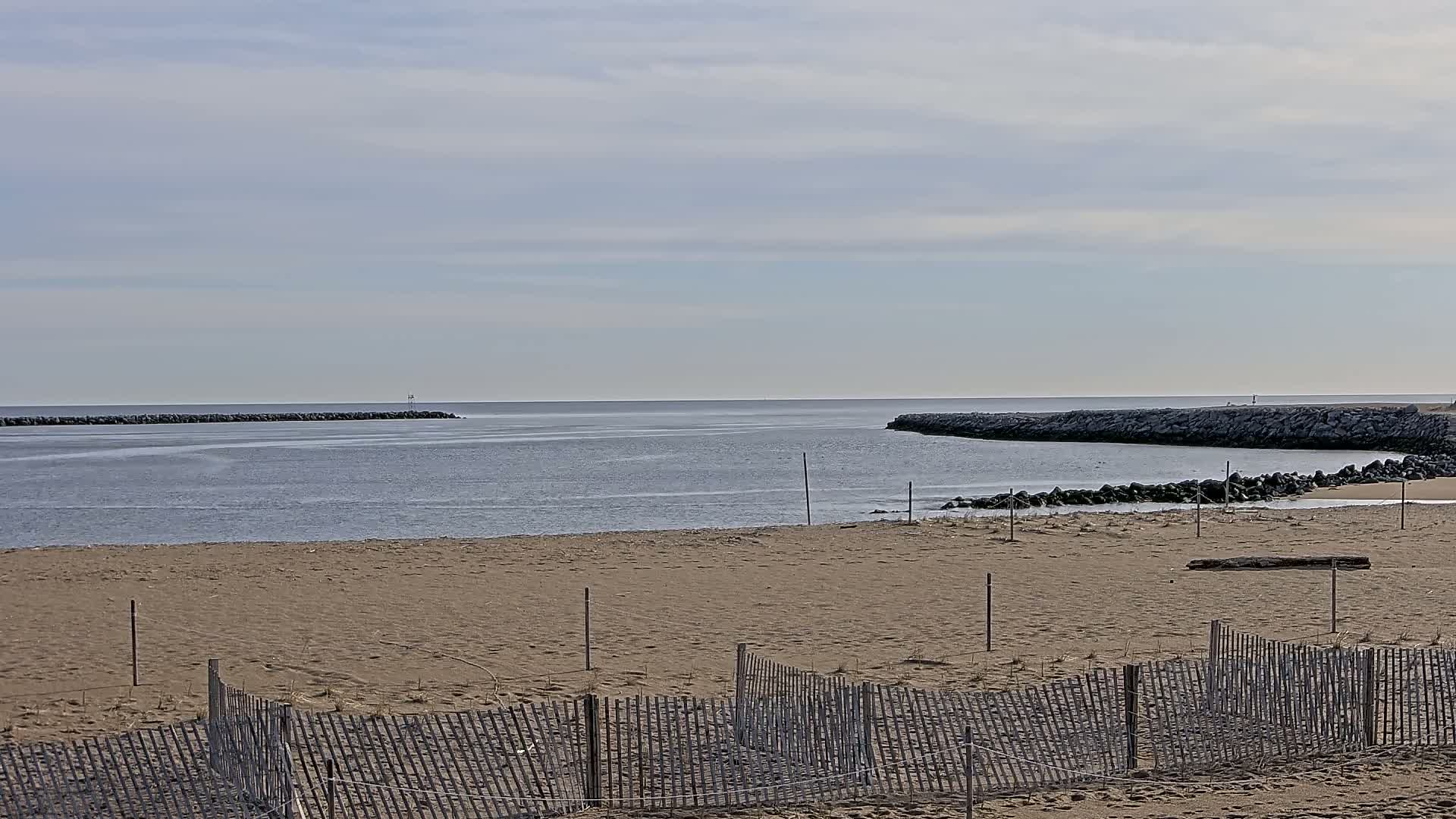 A sandy beach with a wooden fence stretches toward calm, gray water and rock jetties under a mostly cloudy sky.