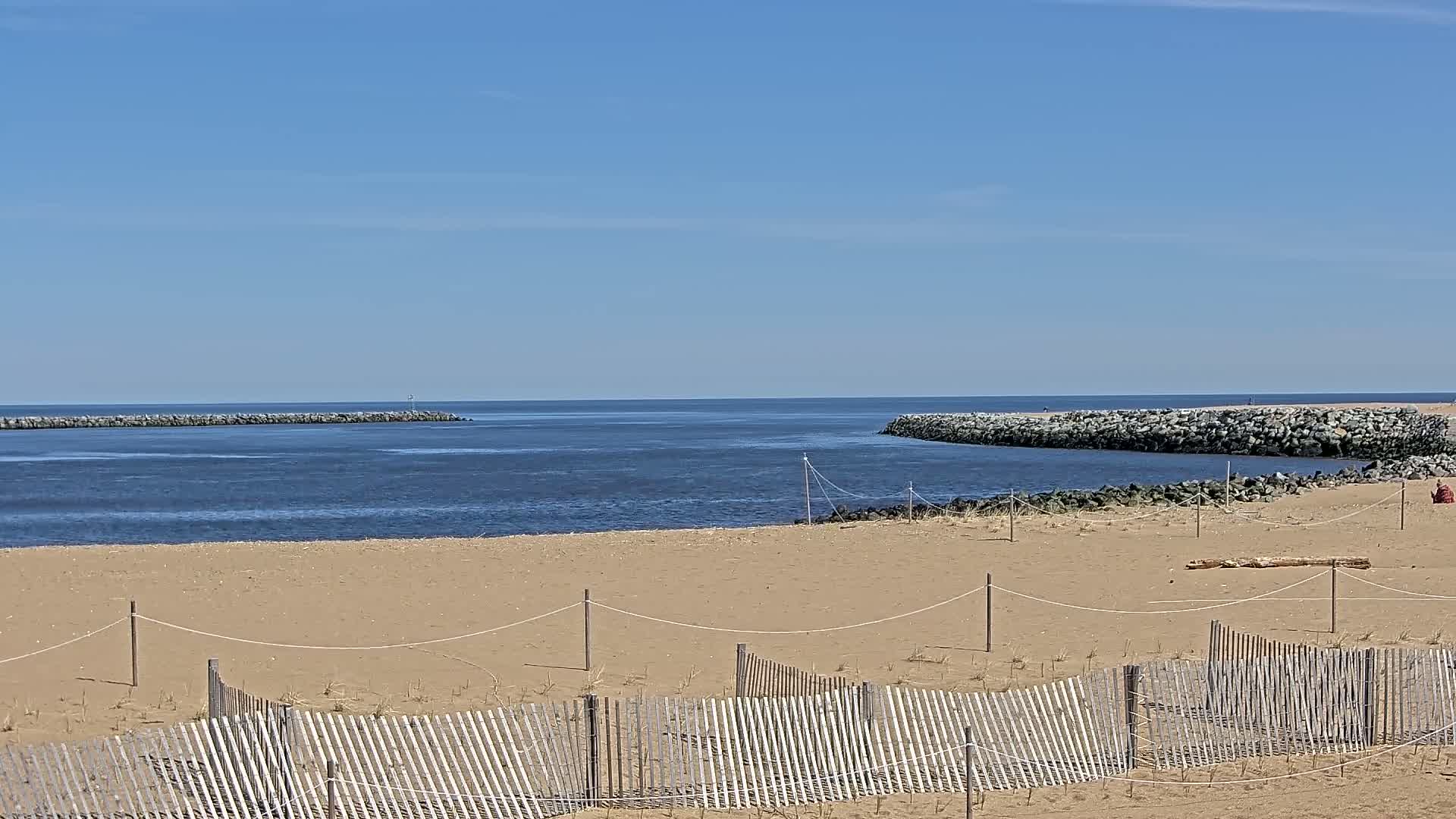 A sandy beach with a wooden fence stretches towards a calm body of water, partially enclosed by rock jetties under a clear blue sky.