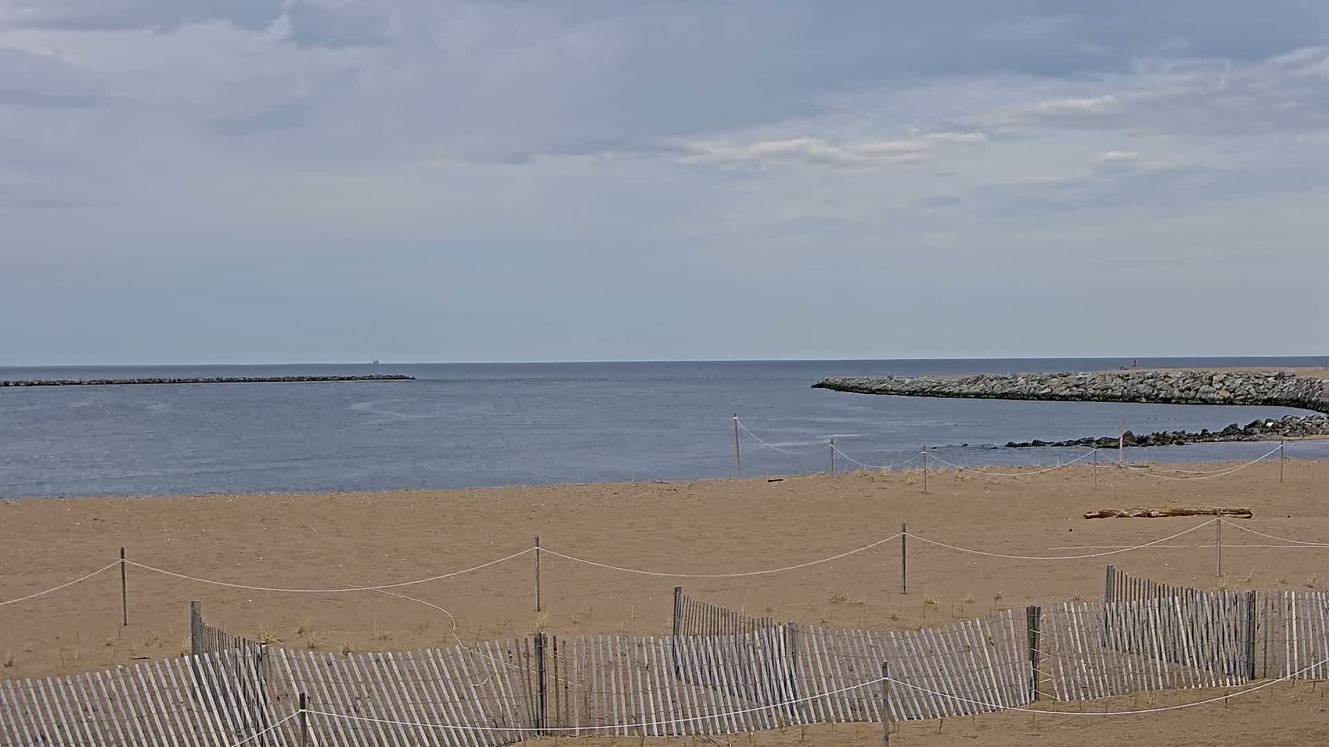 A sandy beach with a wooden fence stretches towards a calm body of water under a mostly cloudy sky,  separated from the open water by rock jetties.