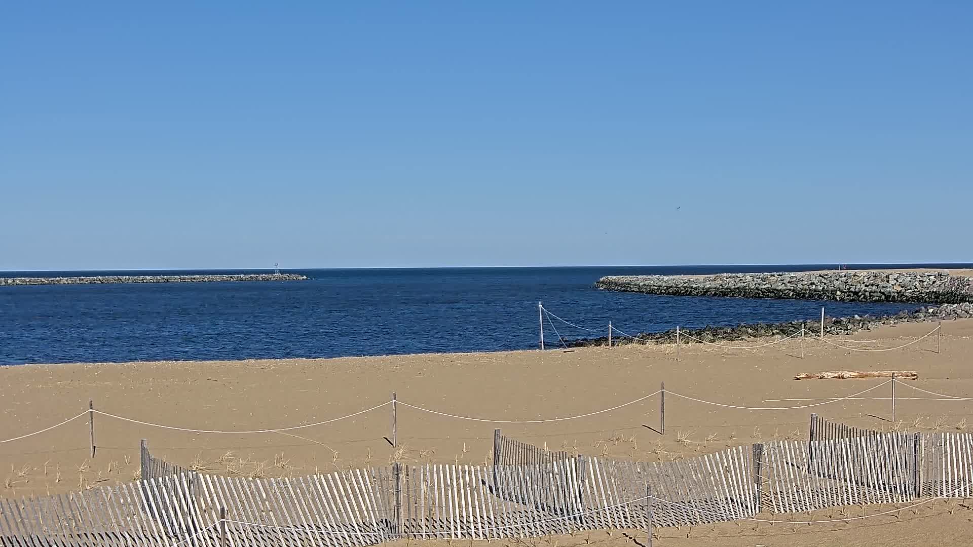 A sandy beach with a wooden fence stretches towards a calm blue ocean, partially enclosed by rock jetties under a clear blue sky.