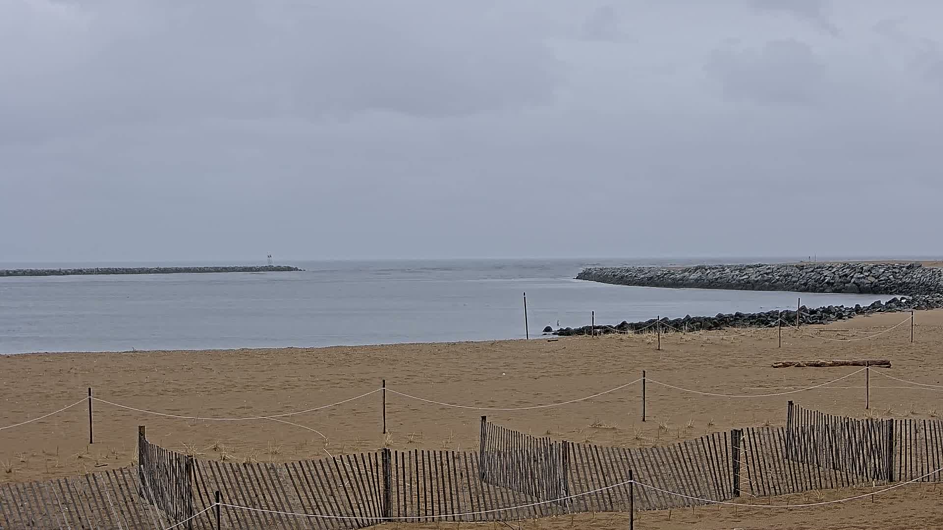 A sandy beach with a low wooden fence stretches toward a calm, gray body of water, bordered by rock jetties under an overcast sky.
