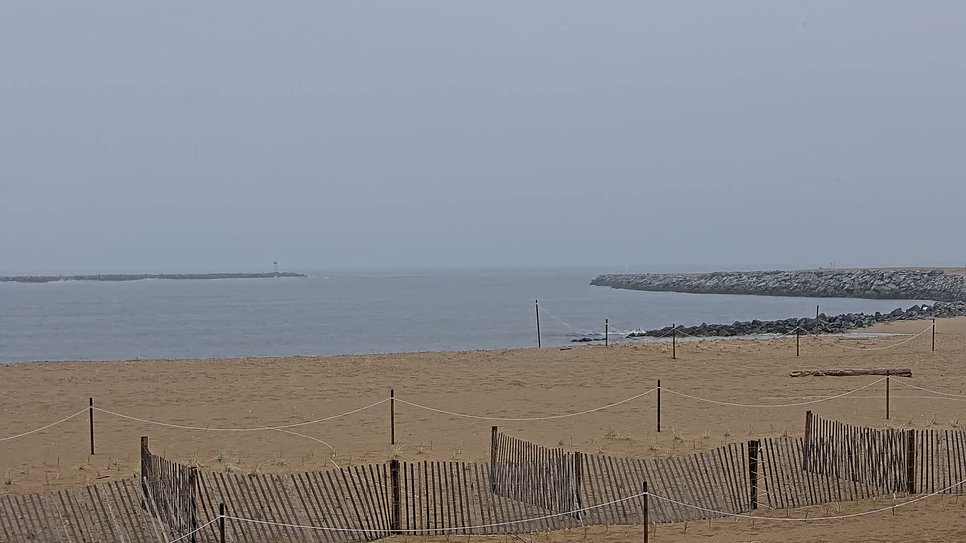 A sandy beach with a wooden fence stretches towards a calm, gray ocean under an overcast sky.