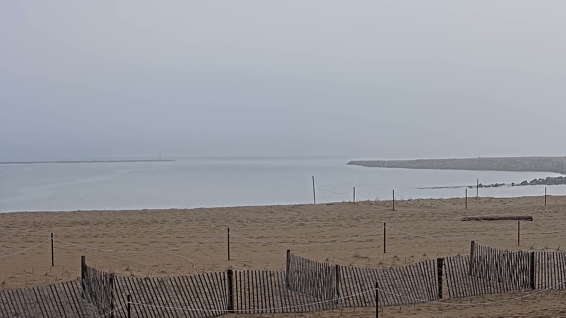 A sandy beach with a wooden fence is visible, overlooking a calm, foggy body of water.