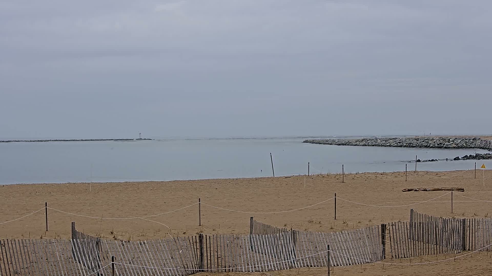 A sandy beach with a wooden fence stretches out to calm, light-grey water under an overcast sky.