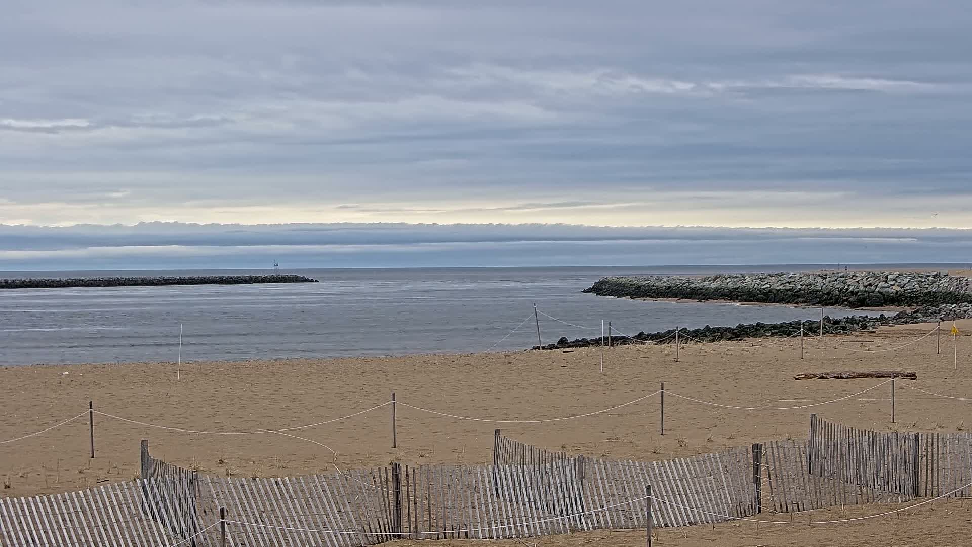 A sandy beach, partially enclosed by a wooden fence, stretches toward a calm ocean under a cloudy sky.