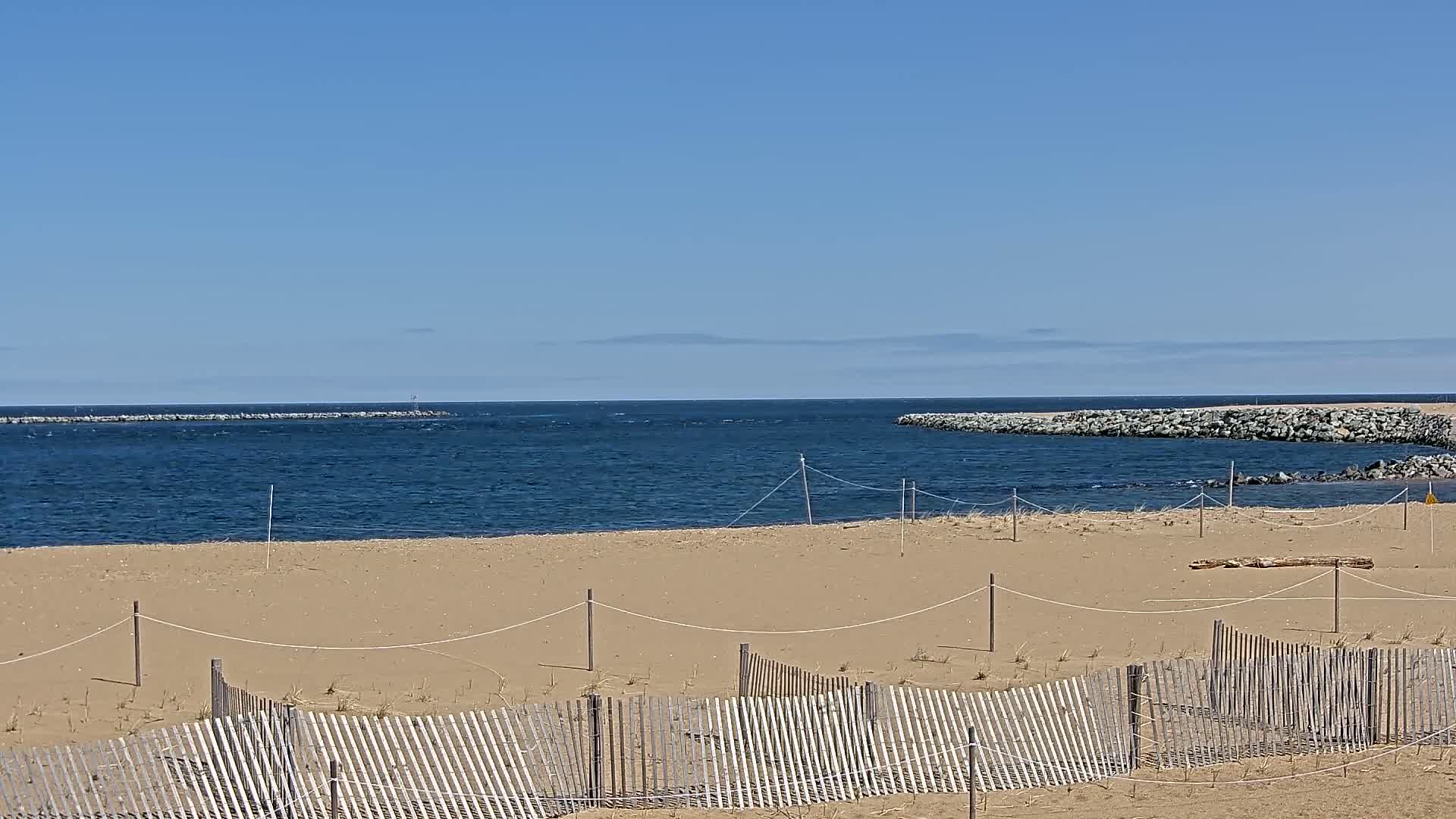 A sandy beach with a wooden fence is seen, overlooking a calm blue ocean partially enclosed by rock jetties under a clear blue sky.