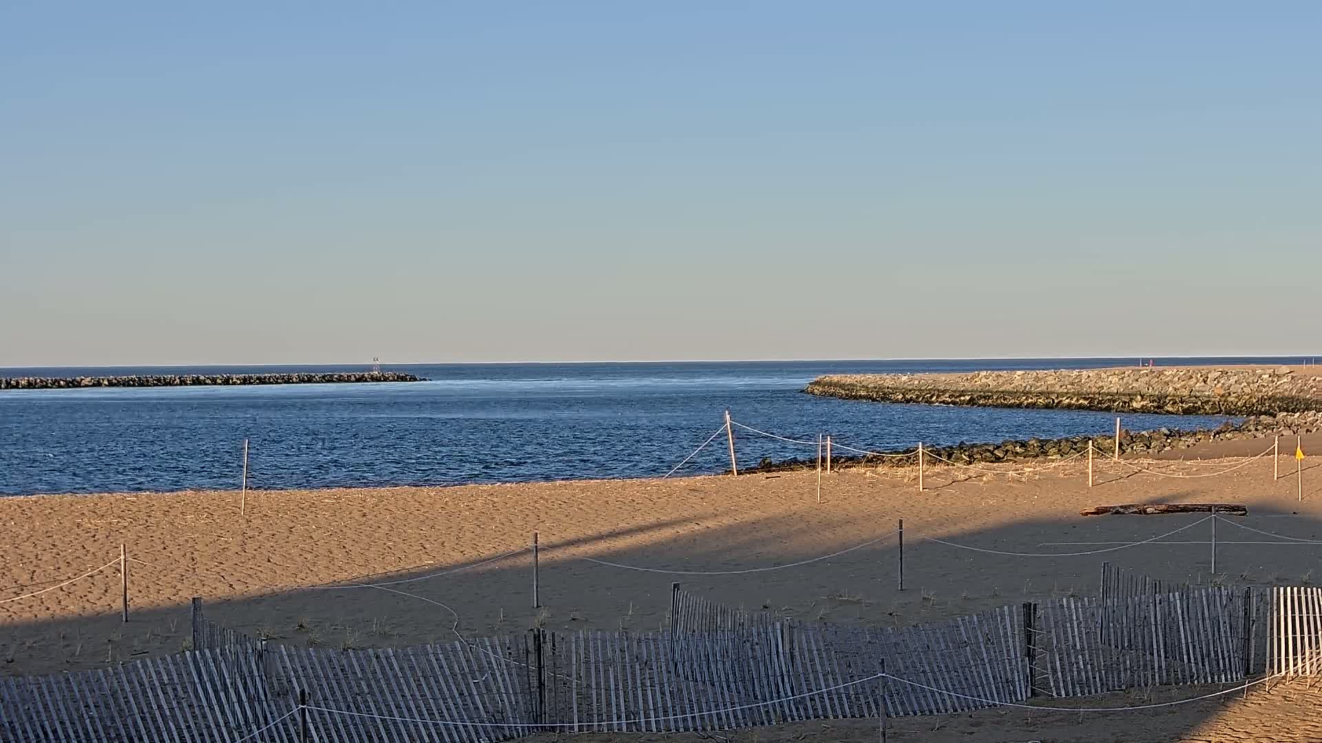 A sandy beach with a wooden fence stretches out to a calm, blue ocean partially enclosed by rock jetties under a clear, bright sky.