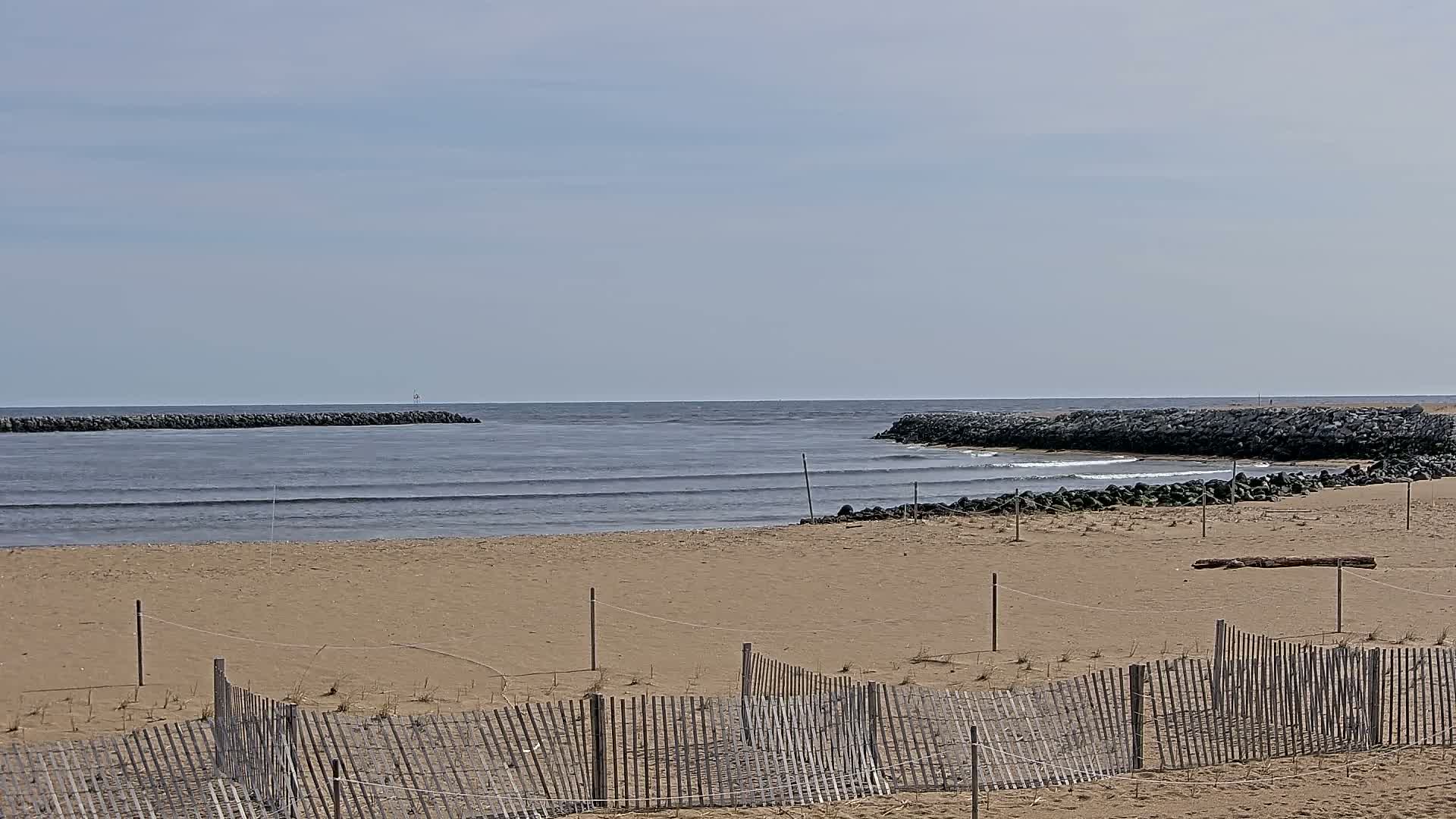 A sandy beach with a wooden fence and small waves on a calm, overcast day, featuring rock jetties extending into the ocean.