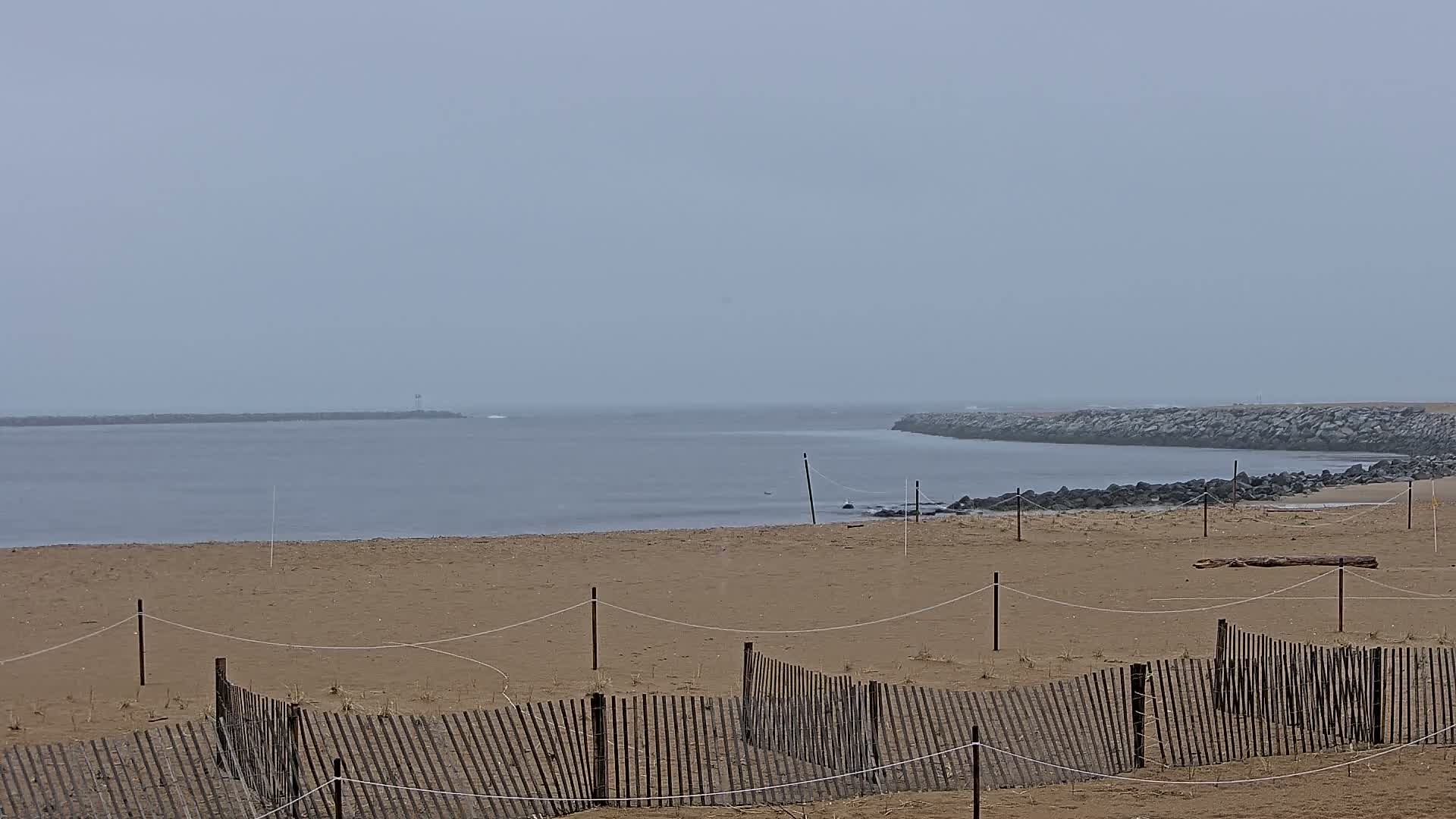 A sandy beach with a wooden fence stretches toward a calm, gray sea under an overcast sky.