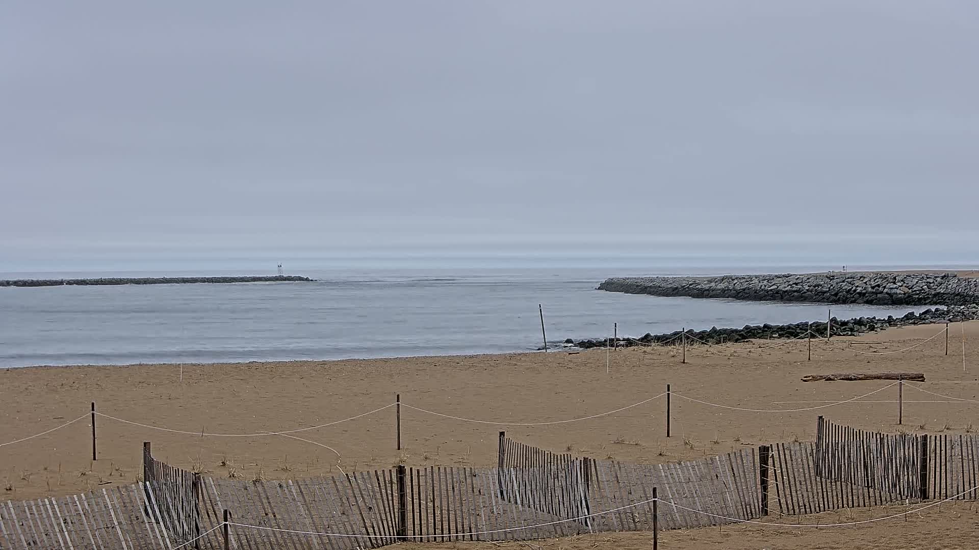 A sandy beach with a wooden fence is seen in the foreground, while a calm, gray ocean with two rock jetties is visible in the background under an overcast sky.