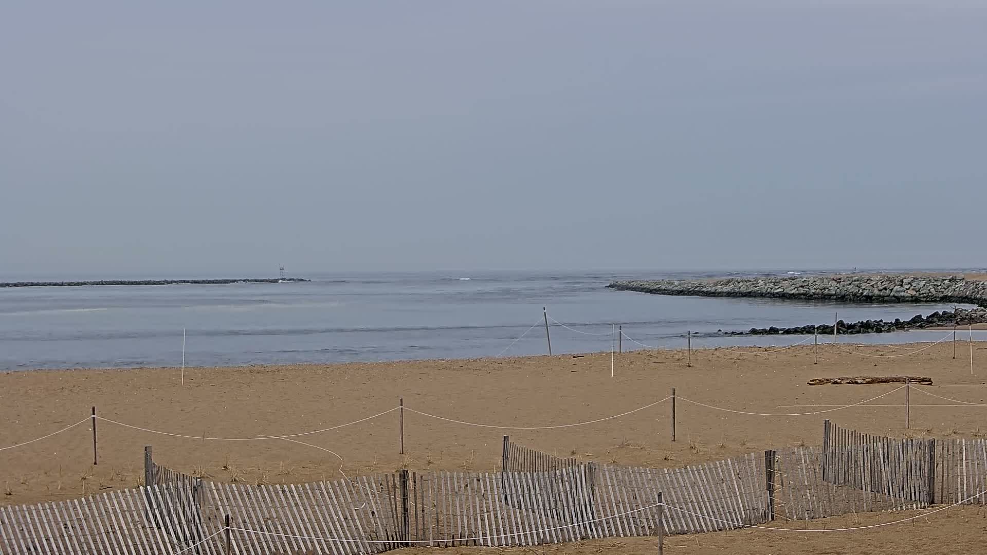 A sandy beach with a wooden fence and thin rope lines stretches towards a calm, gray-toned ocean inlet bordered by rock jetties under an overcast sky.