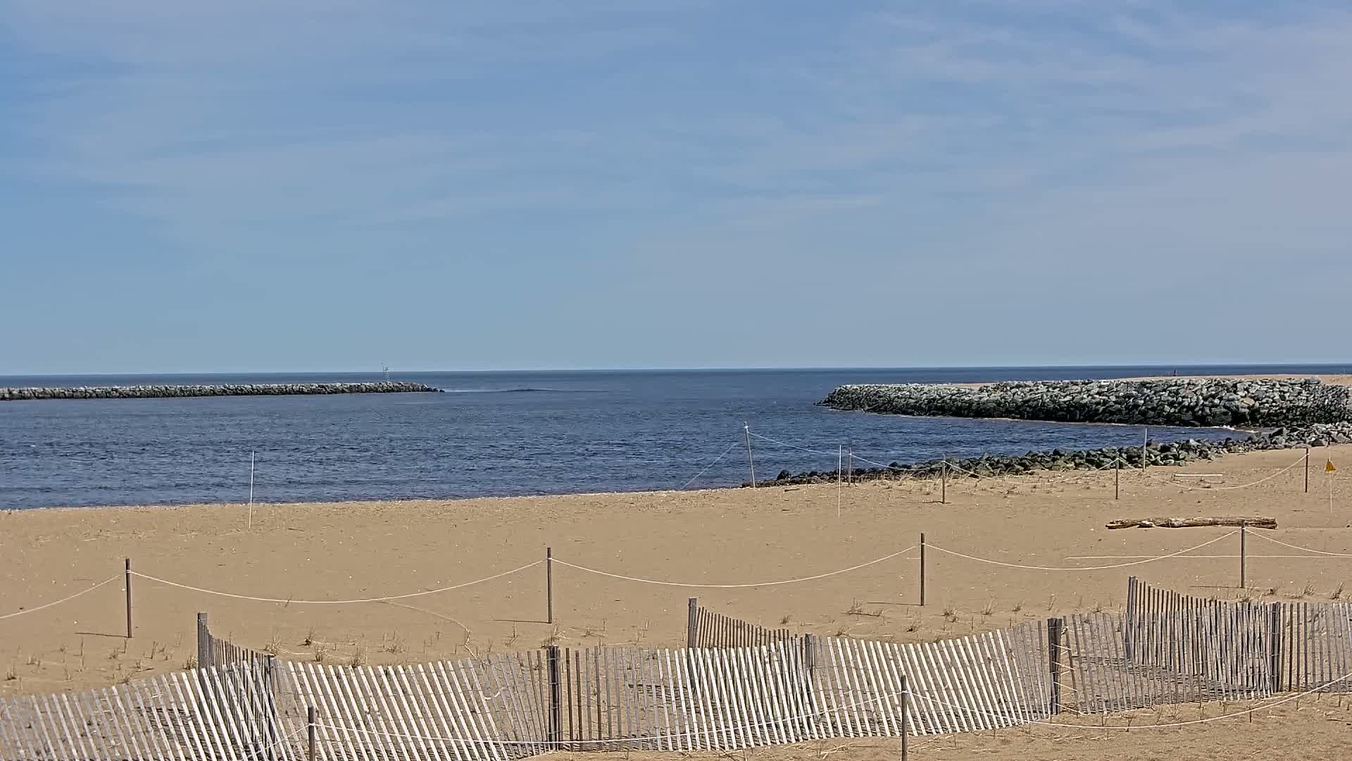 A sandy beach with a wooden fence is shown, overlooking a calm body of water with rock jetties under a mostly clear, sunny sky.