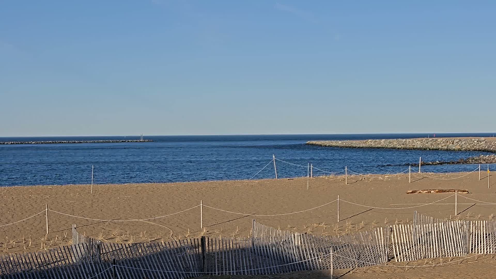A sandy beach with a wooden fence, ropes, and a log, overlooks a calm blue ocean inlet bordered by rock jetties under a clear blue sky.
