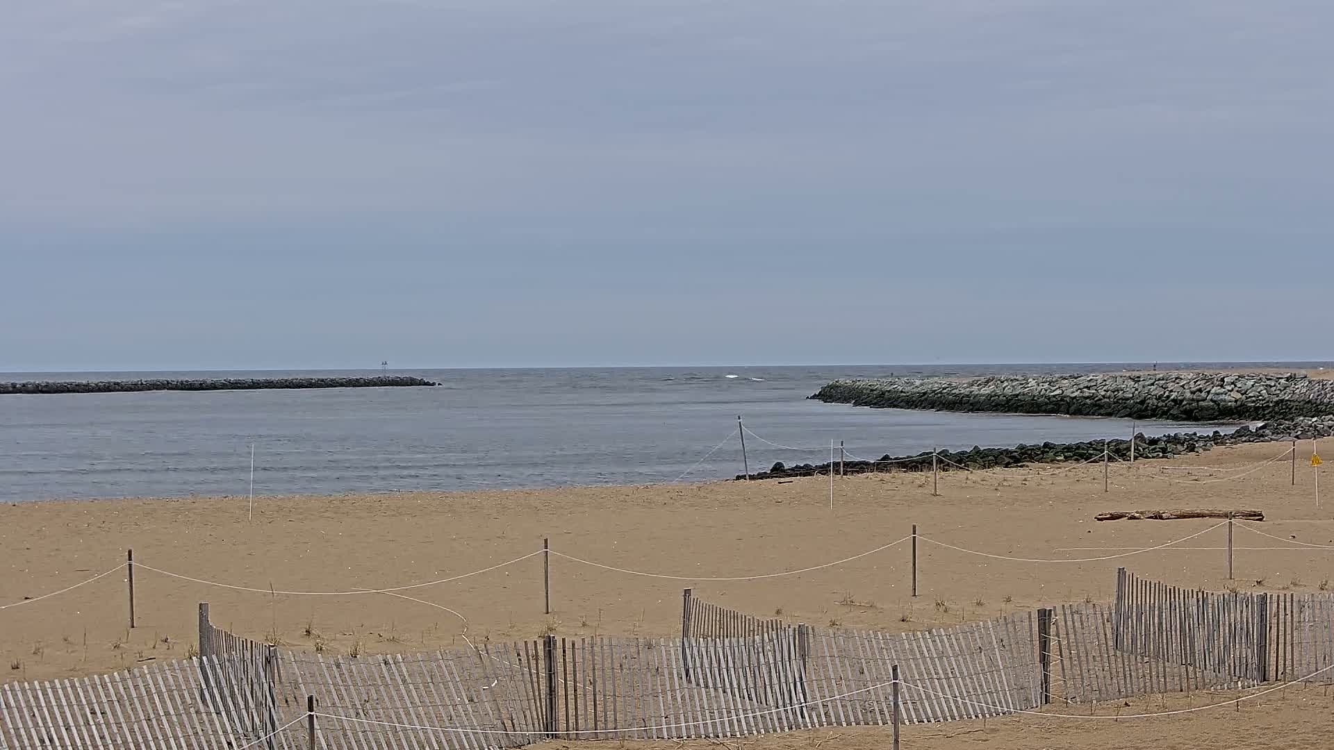 A sandy beach with a wooden fence is seen, encompassing a calm, gray ocean with rock jetties on either side under an overcast sky.