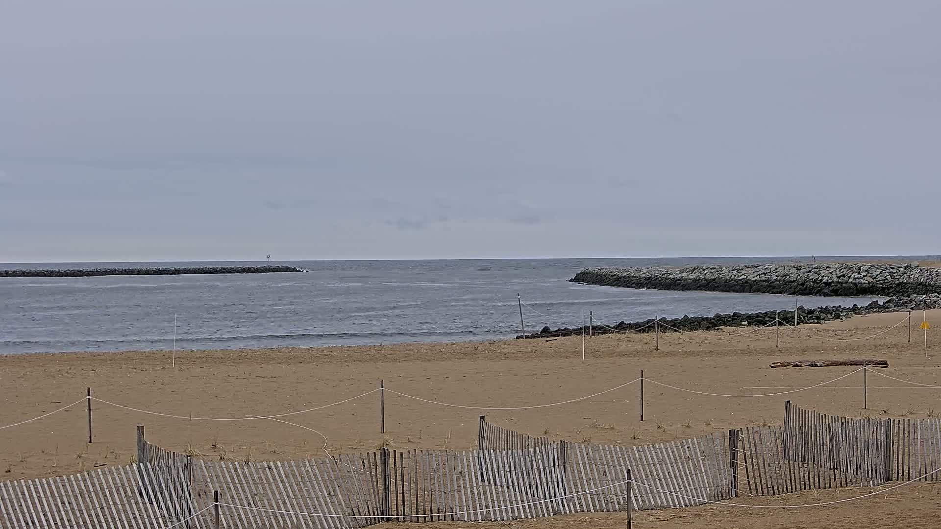 A sandy beach with a wooden fence is partially visible, separating it from calm, grey water and rock jetties under an overcast sky.