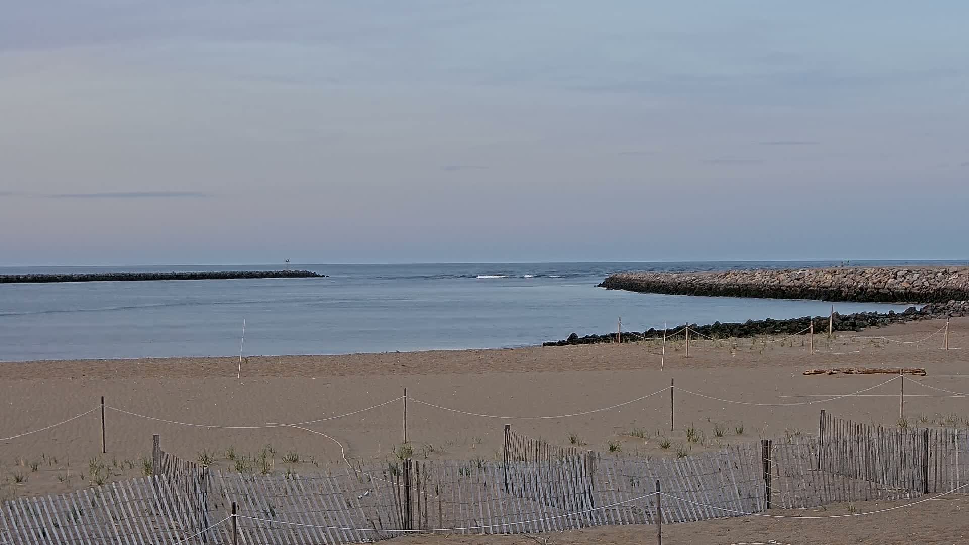 A sandy beach with a wooden fence is seen, leading to a calm body of water bordered by stone jetties under a cloudy sky.