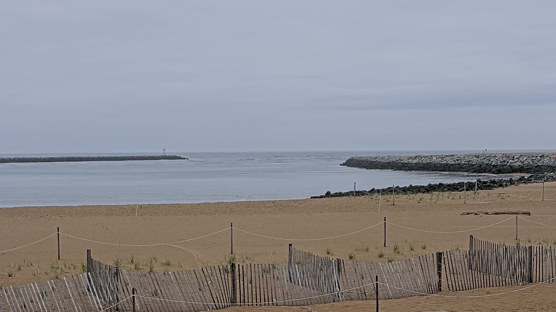 A sandy beach with a low wooden fence is visible, overlooking calm water and rock jetties under an overcast sky.