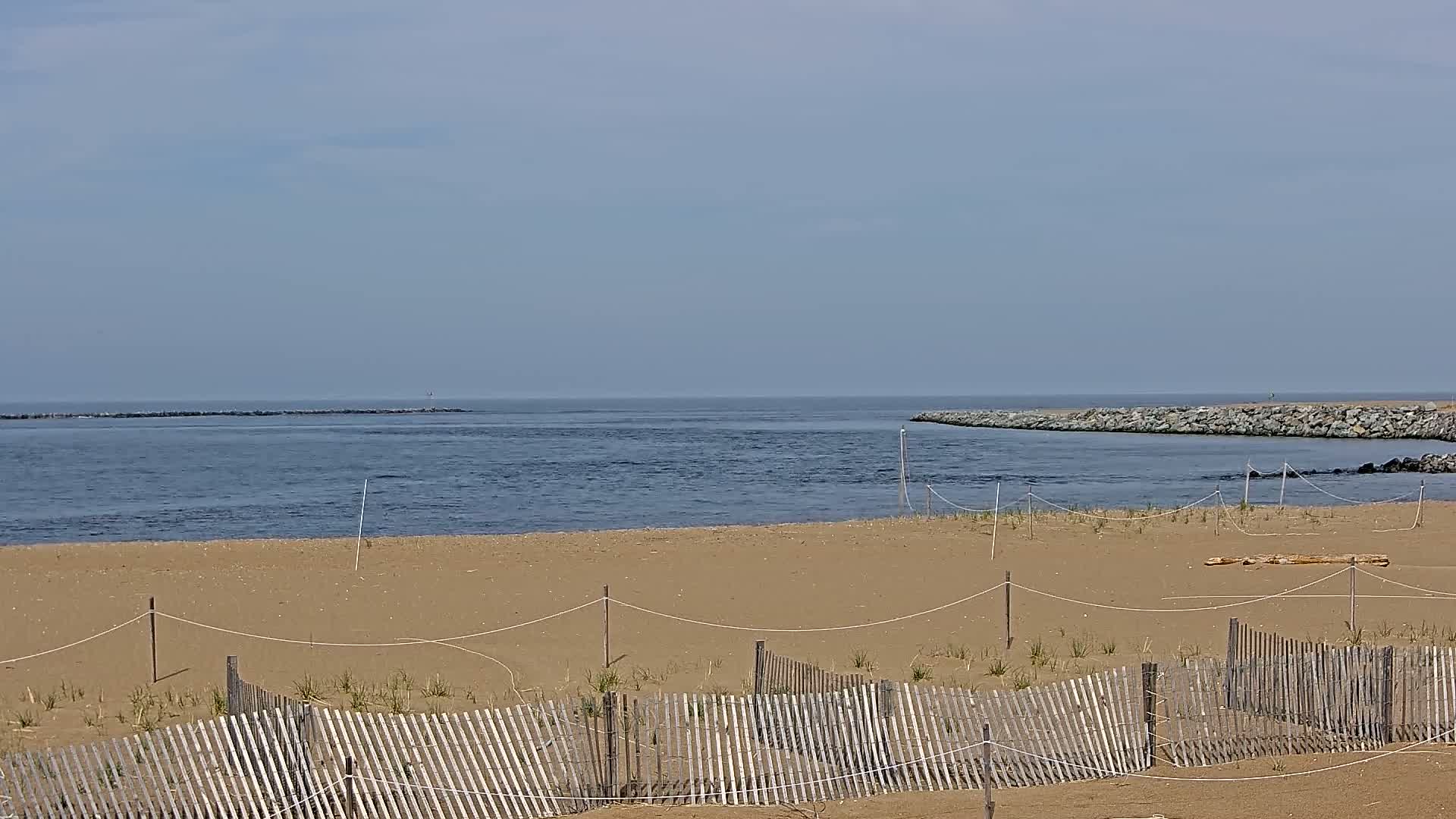A sandy beach with a wooden fence stretches toward a calm body of water, partially enclosed by rock jetties under a clear sky.