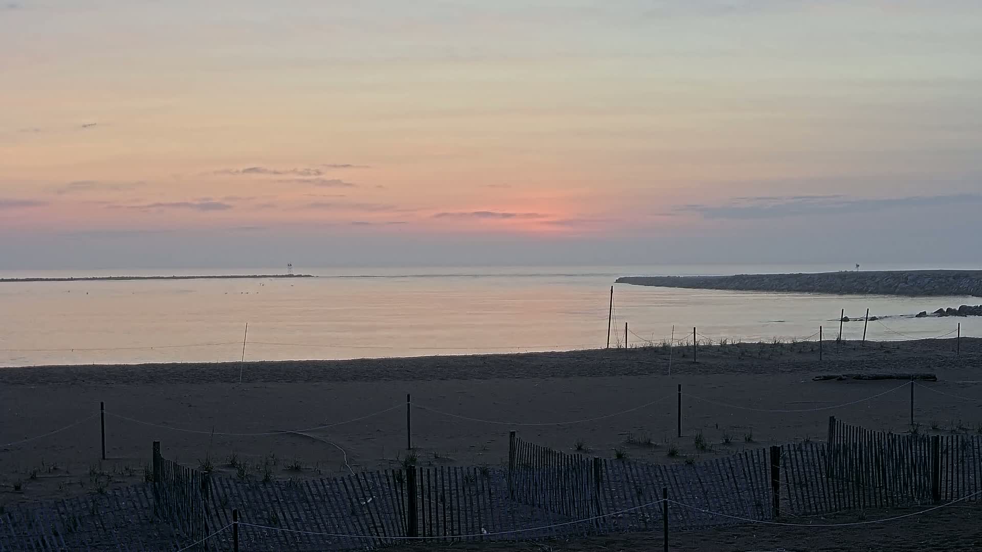 A calm, pinkish sunrise over a body of water, viewed from a sandy beach with a low wooden fence in the foreground.