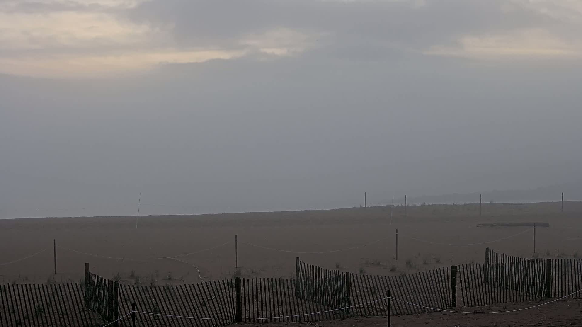 A foggy beach scene shows a sandy expanse with a low wooden fence and thin posts stretching across it.