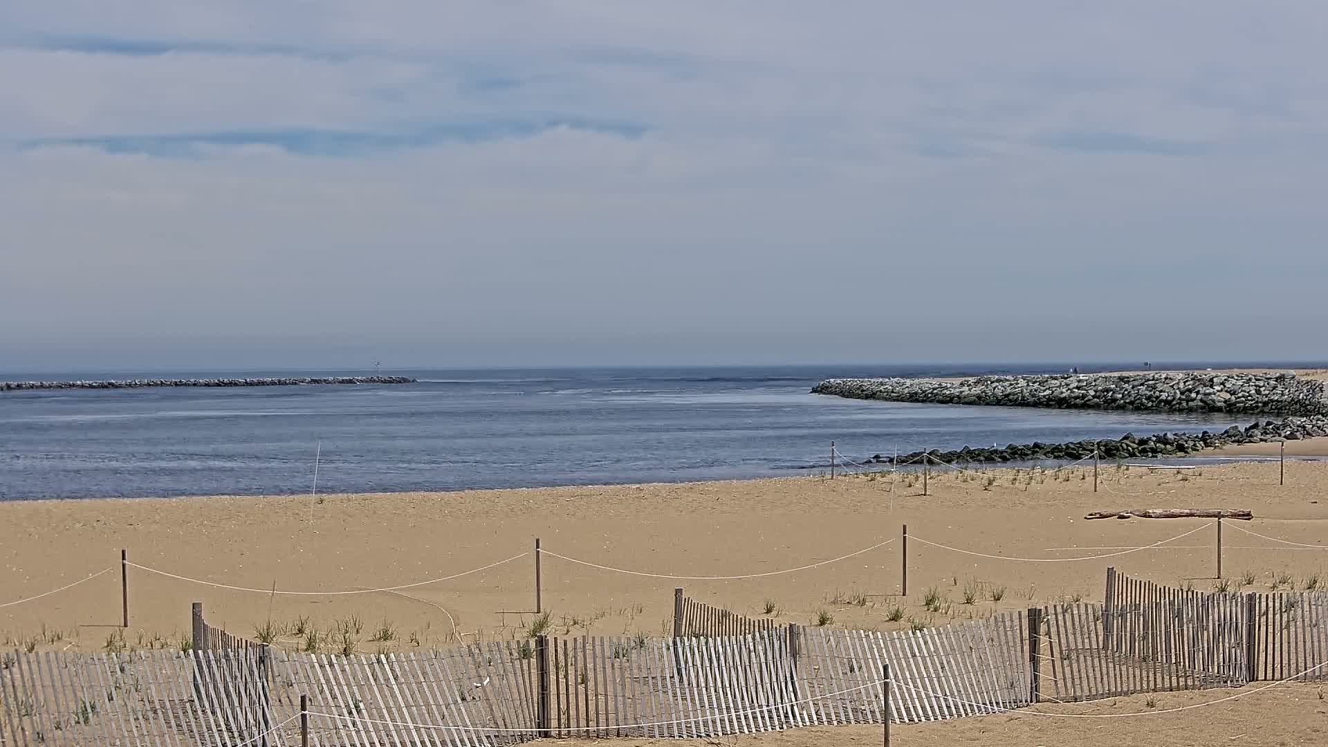 A sandy beach with a wooden fence stretches toward a calm inlet bordered by rock jetties under a partly cloudy sky.