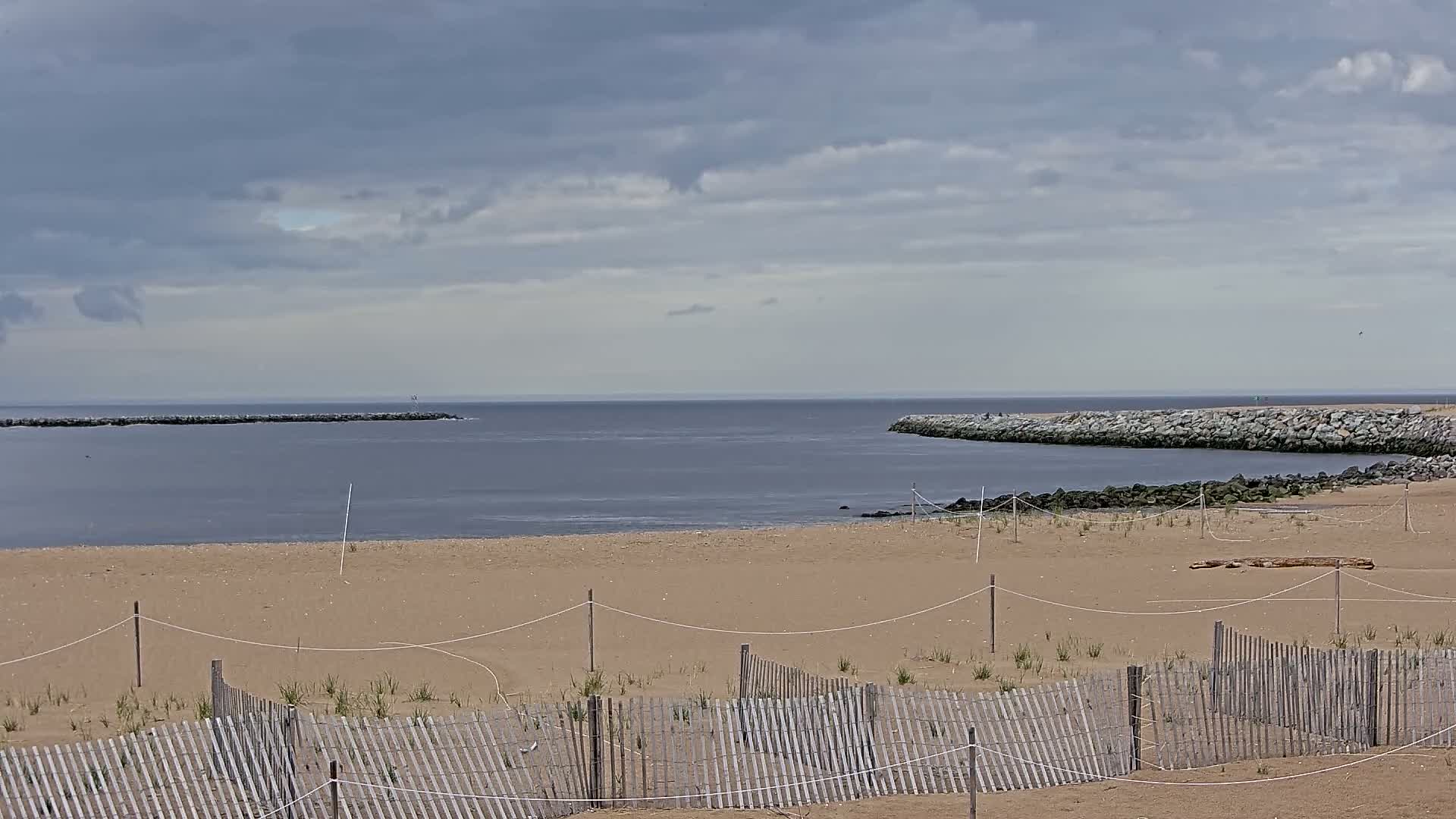 A sandy beach with a wooden fence is partially visible, looking out to calm, gray water partially enclosed by rock jetties under a cloudy sky.