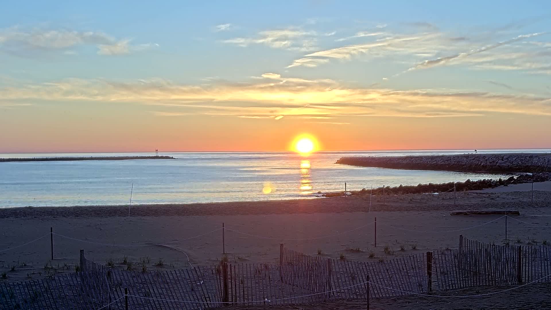 A sandy beach with a low wooden fence is seen at sunset, with the sun rising over calm water between two jetties.