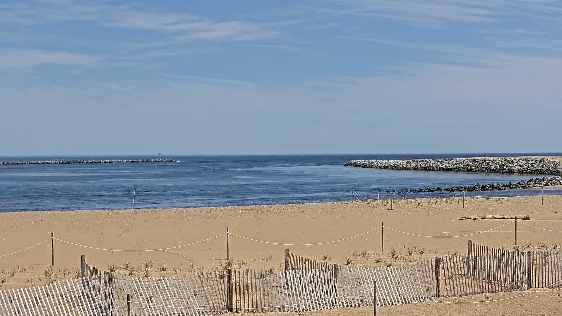 A sandy beach with a wooden fence stretches towards a calm, blue inlet bordered by rock jetties under a mostly sunny sky.