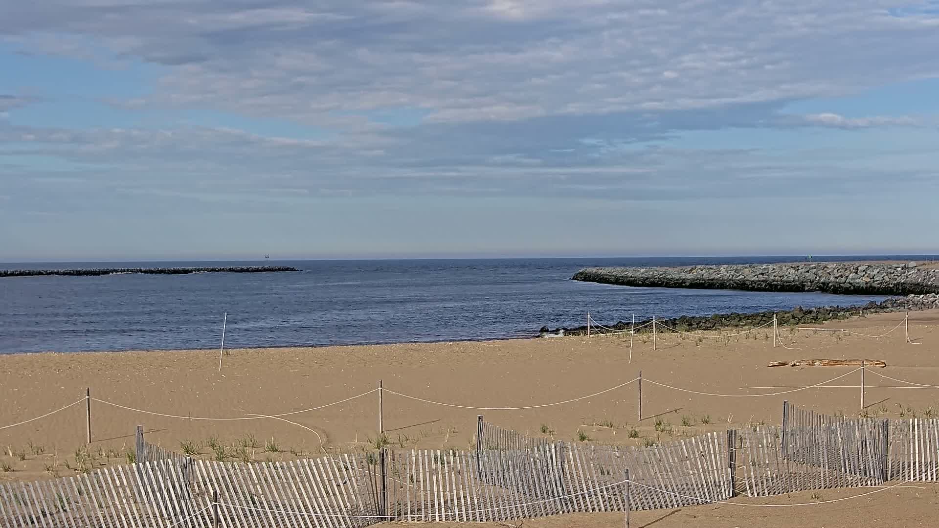 A sandy beach with a low wooden fence stretches toward a calm, blue ocean inlet bordered by dark rock jetties under a partly cloudy sky.