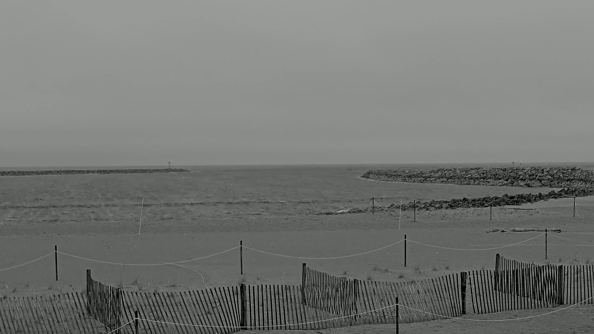 A grayscale image shows a beach with a simple wooden fence, under an overcast sky, with calm waves and rock jetties visible in the distance.