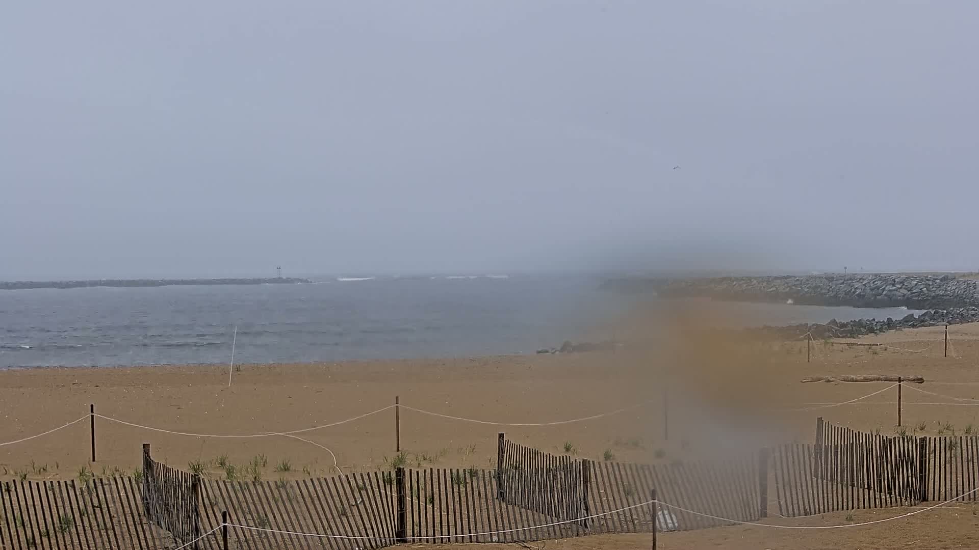 A hazy, overcast day at a beach shows a sandy shore with a wooden fence and calm waves in the distance.