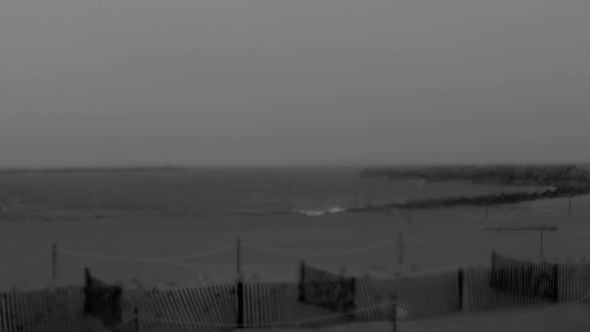 A foggy, overcast day at the beach shows a choppy ocean with a breakwater and a section of beach with wooden fences in the foreground.