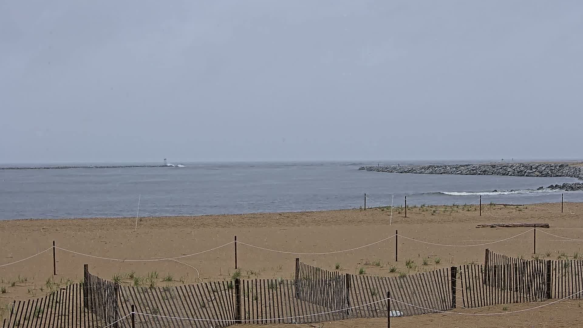 A sandy beach with a low wooden fence is seen, overlooking a calm, gray ocean under an overcast sky.
