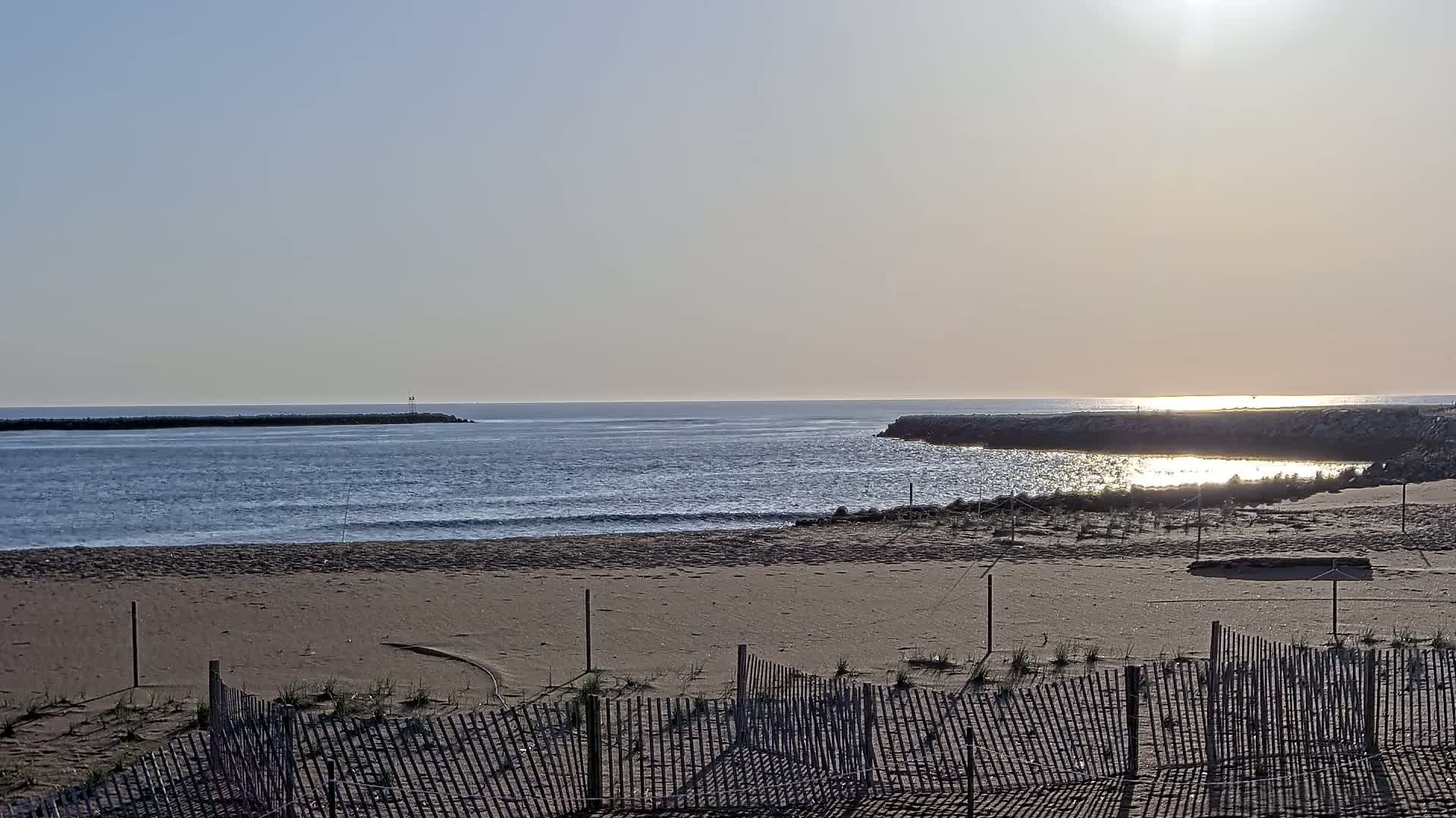 A sandy beach with a wooden fence is shown, overlooking calm ocean water with a breakwater on either side, under a bright, sunny sky.