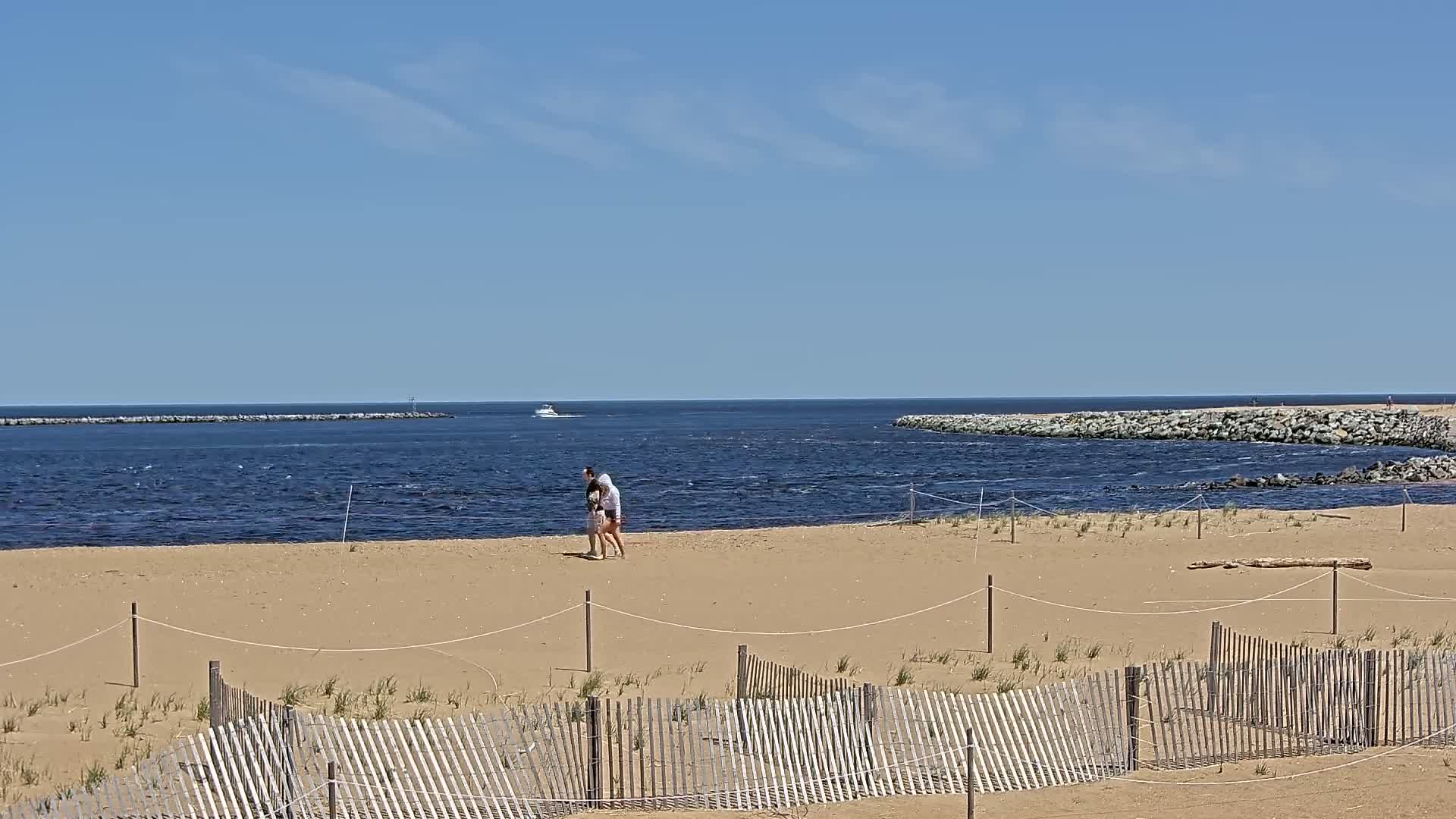 A sandy beach with a wooden fence, a couple walking, and a calm ocean with a boat in the distance under a clear, sunny sky.