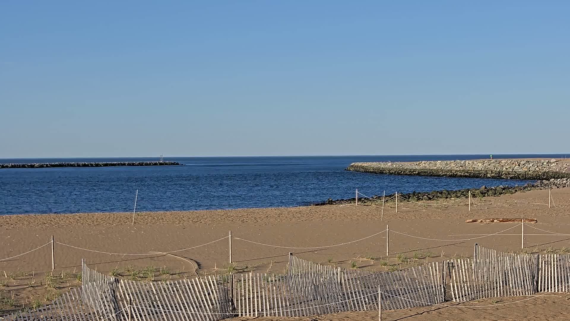 A sandy beach with a wooden fence is bordered by calm blue water and rock jetties under a clear blue sky.