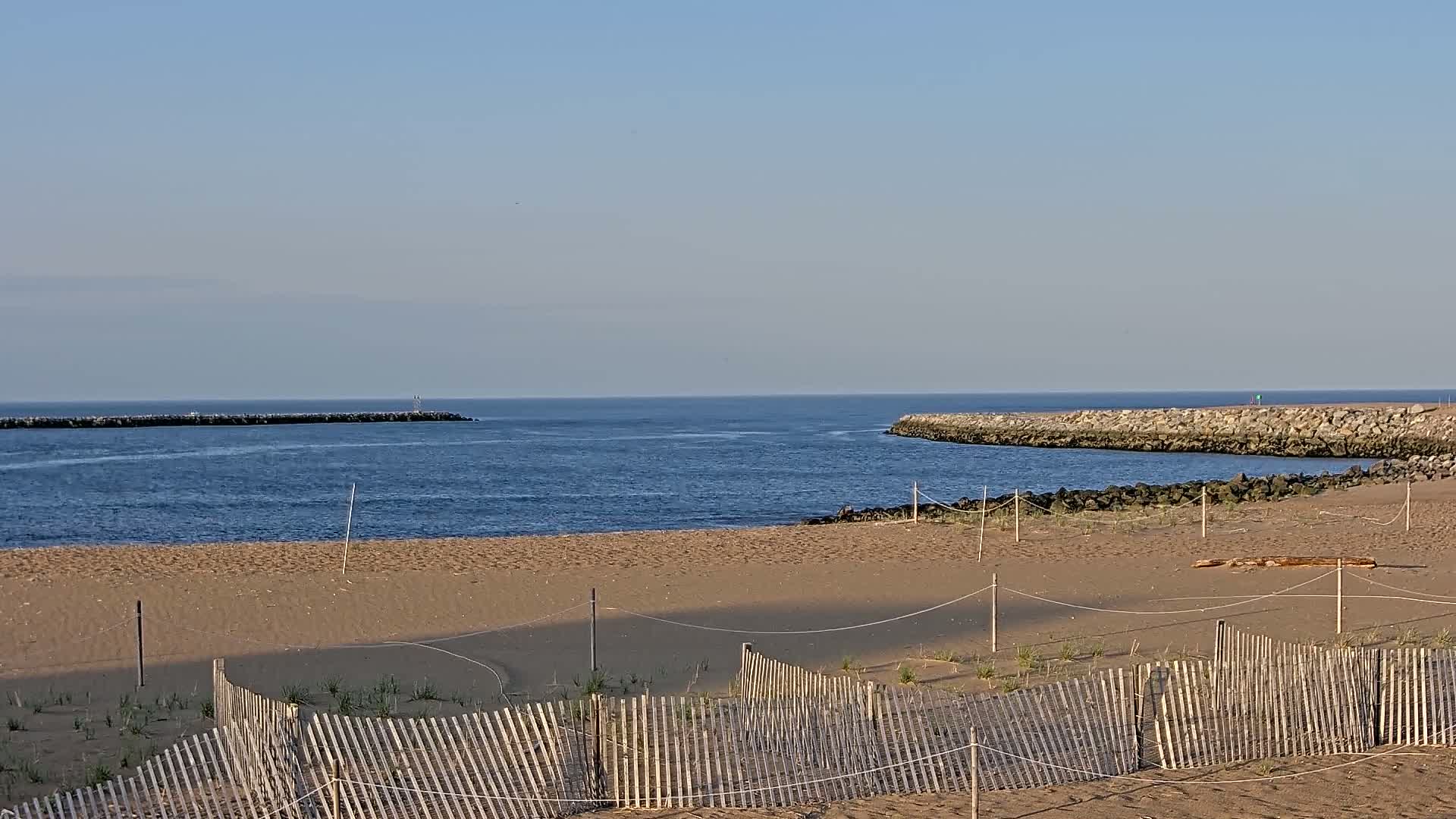 A sandy beach with a wooden fence stretches towards a calm, blue ocean inlet bordered by rock jetties under a clear sky.