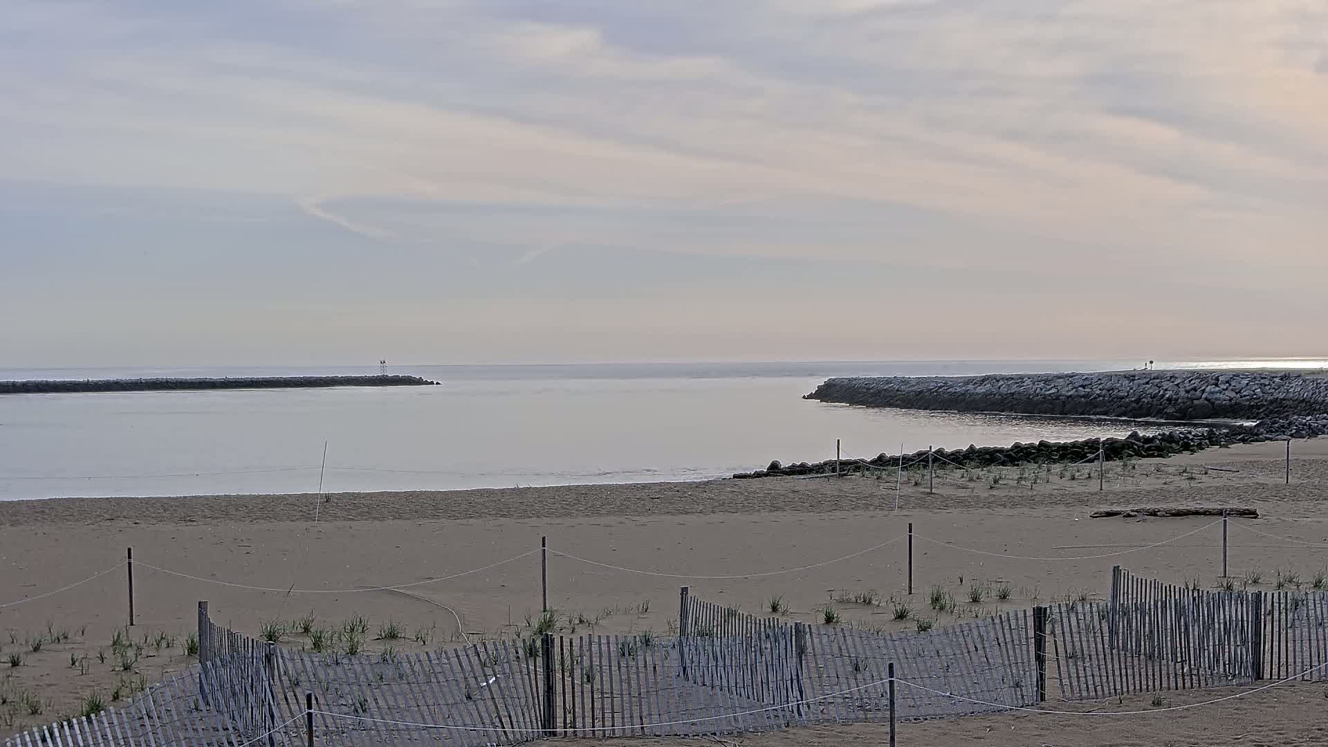 A sandy beach with a low wooden fence stretches to a calm, light-grey ocean inlet bordered by rock jetties under a cloudy sky.