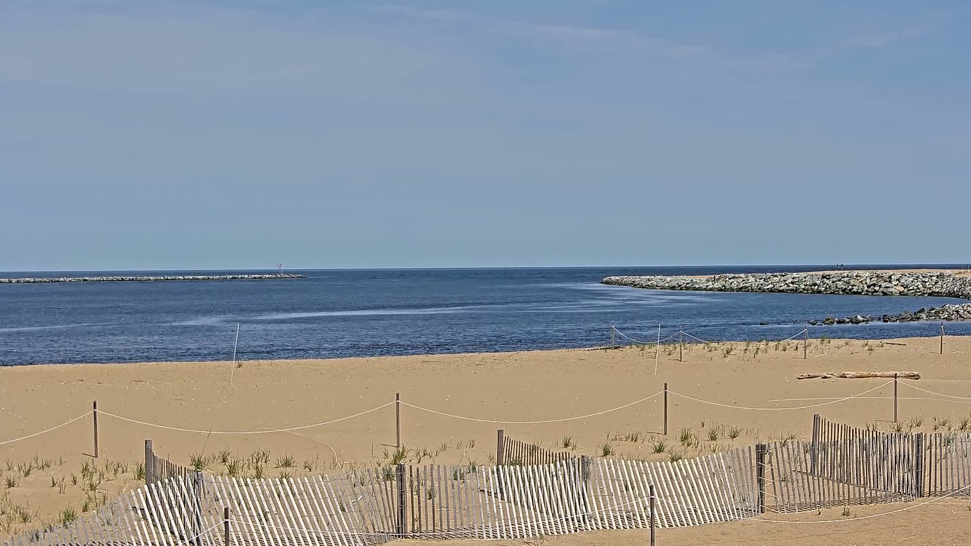 A sandy beach with a wooden fence is shown, leading to a calm body of water with rock jetties on either side under a clear, bright sky.