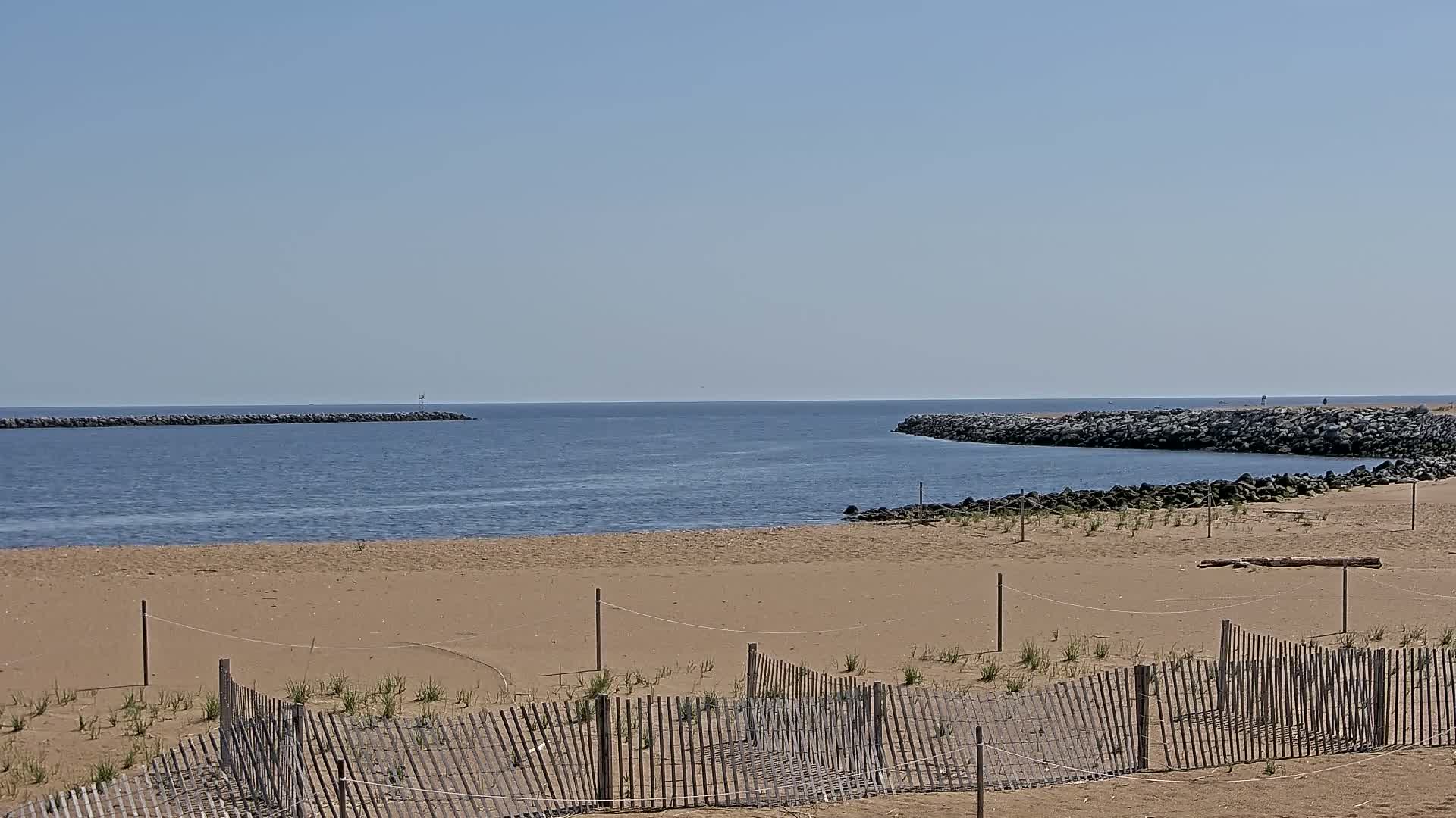 A sandy beach with a wooden fence and rock jetties on either side of a calm, blue body of water under a clear sky.