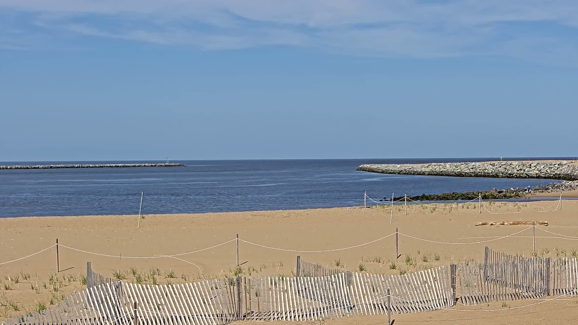A sandy beach with a low wooden fence is seen, under a mostly sunny sky, overlooking a calm inlet bordered by rock jetties.