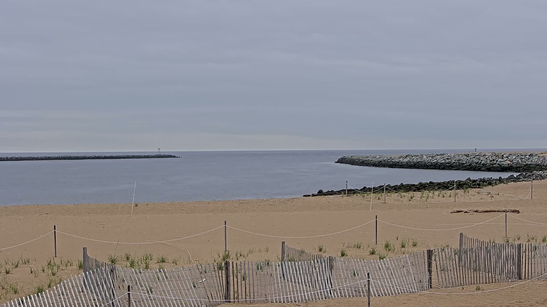 A sandy beach with a wooden fence and sparse vegetation stretches toward a calm body of water bordered by rock jetties under an overcast sky.