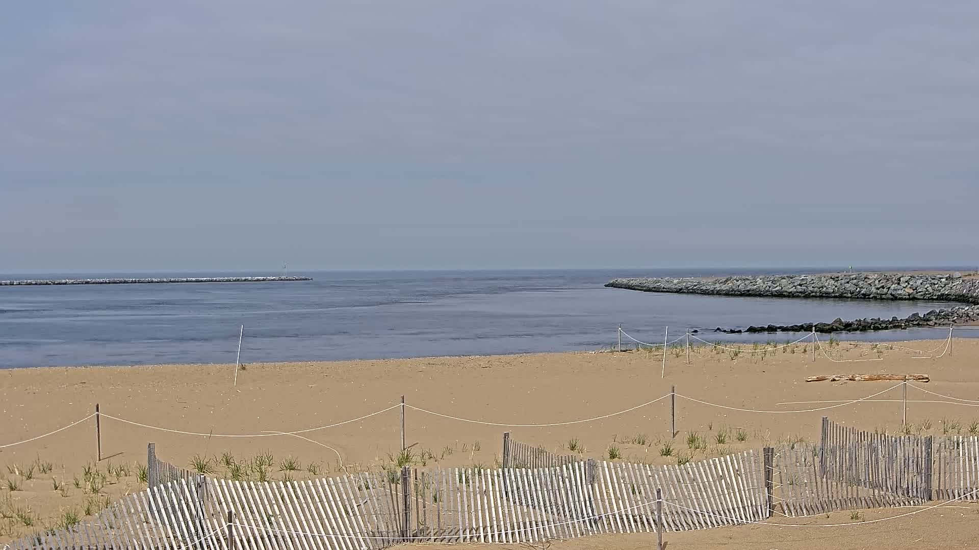 A sandy beach with a low wooden fence stretches towards a calm body of water, partially enclosed by rock jetties under a hazy sky.