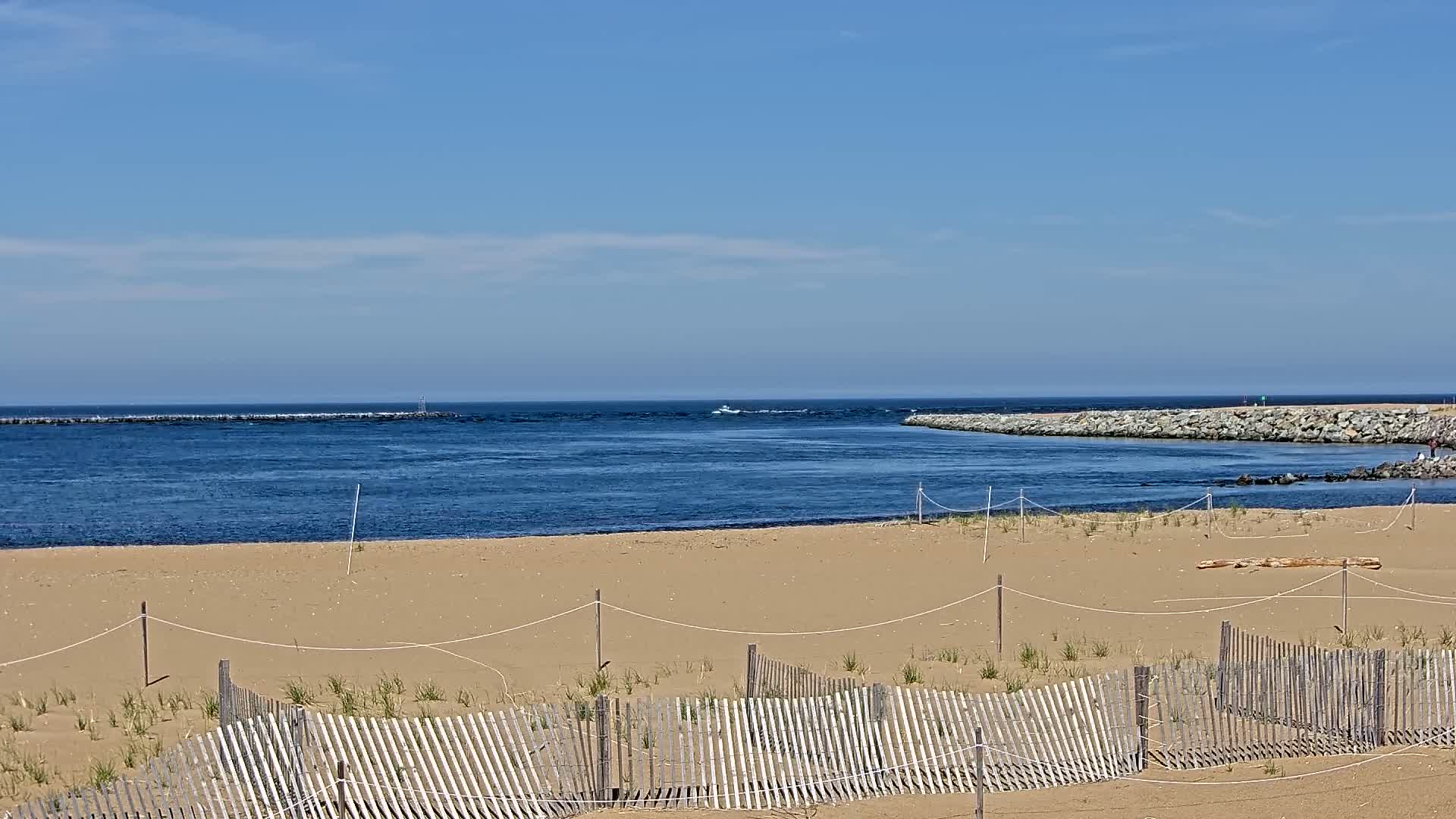 A sandy beach with a wooden fence is shown, overlooking a calm ocean inlet with a small boat visible in the distance under a clear blue sky.