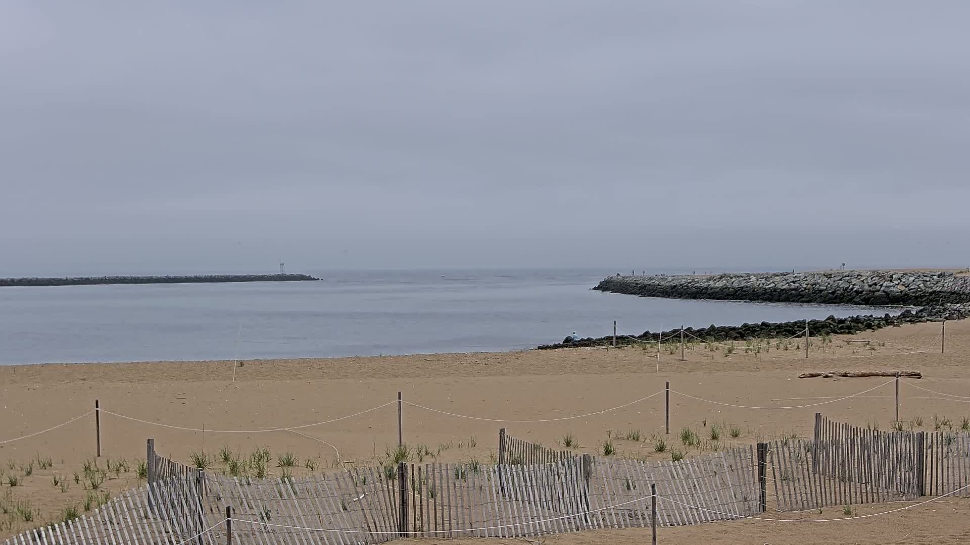 A sandy beach with a wooden fence is situated between two rock jetties on a calm, overcast day.