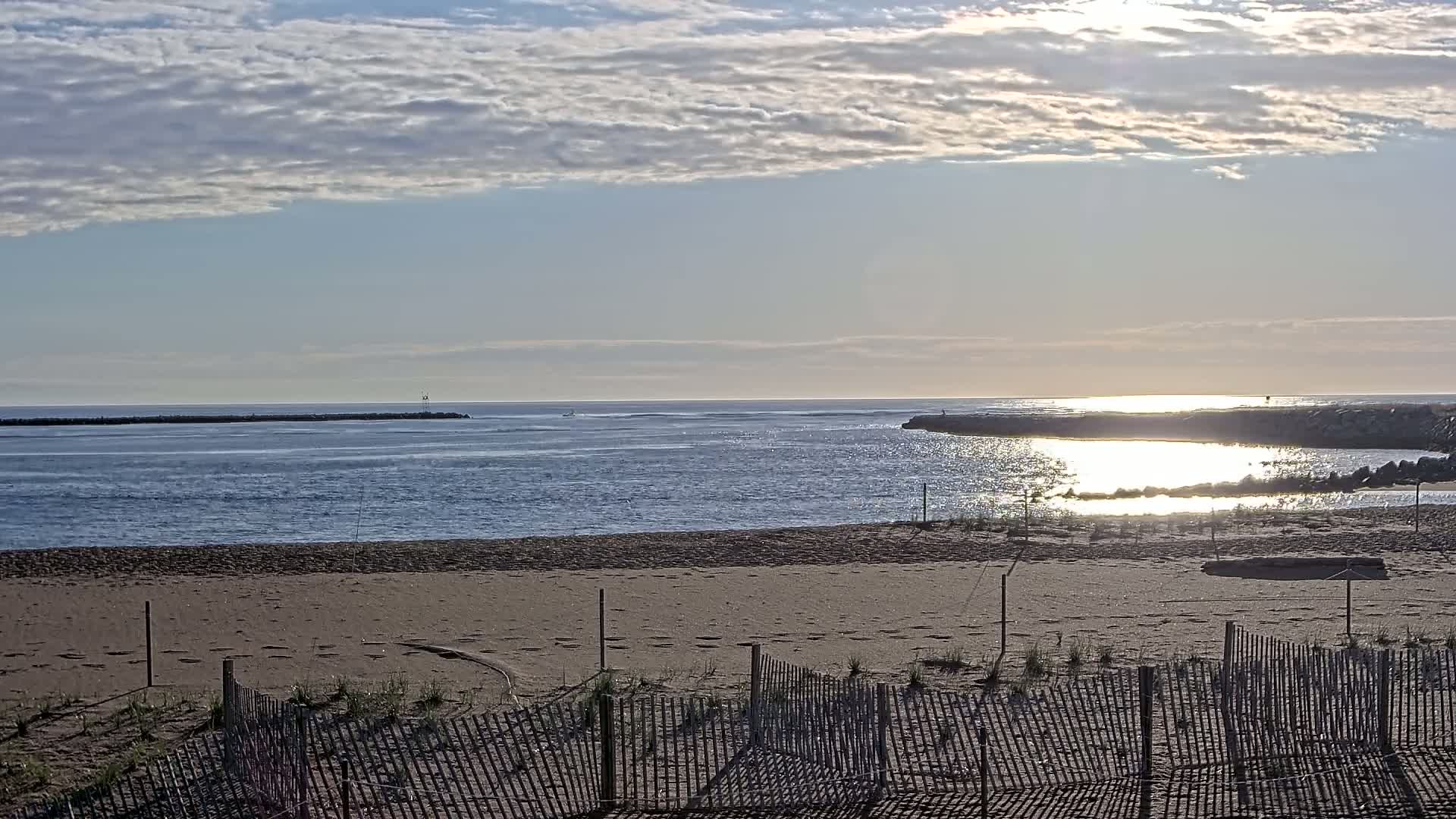 A sandy beach with a wooden fence stretches towards a calm ocean under a partly cloudy sky, with the sun reflecting off the water.