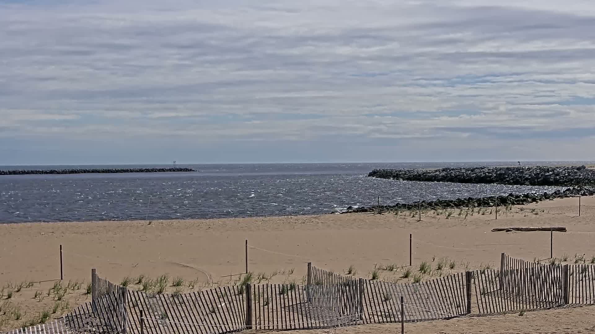 A sandy beach with a wooden fence stretches towards a choppy, gray-blue ocean under a mostly cloudy sky.
